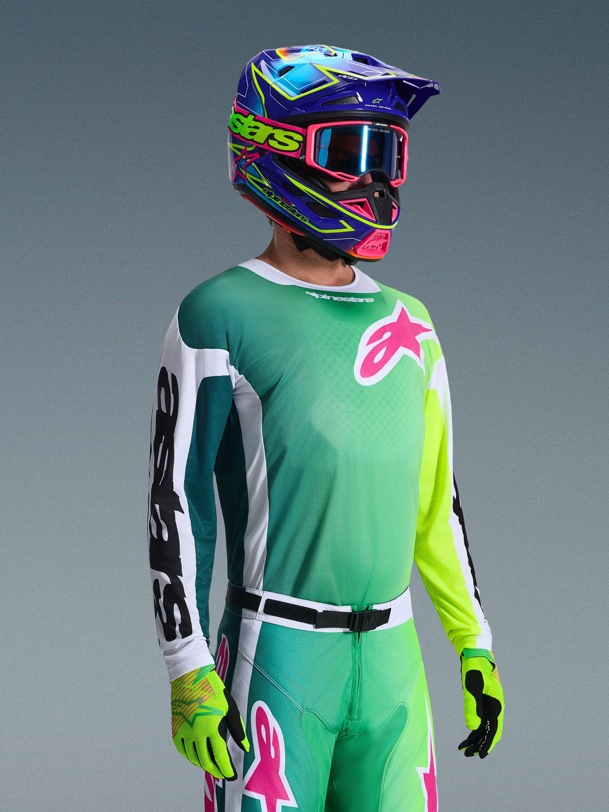 Alpinestars 2026 Racer Portl Jersey, motocross shirt, groen, wit en zwart, gedragen door een rijder met bijpassende groene broek, fluorescerende handschoenen en een opvallende blauwe en paarse helm met roze crossbril, voorzien van een roze Astars-logo op de borst