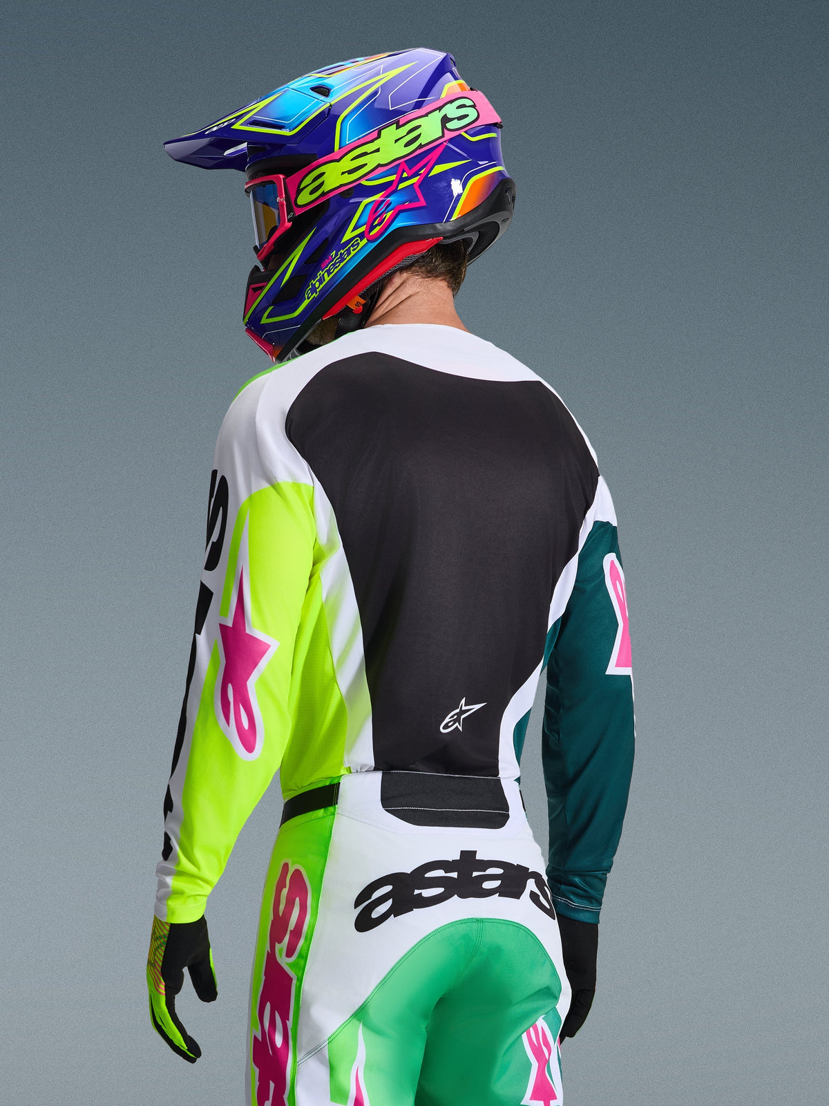 Alpinestars 2026 Racer Portl Jersey, motorcrossrijder van achteren gezien in het groen, wit en zwart met roze sterlogo's, gecombineerd met bijpassende broek en een kleurrijke blauw-paarse helm, gecoördineerde race-uitrusting voor off-road prestaties.