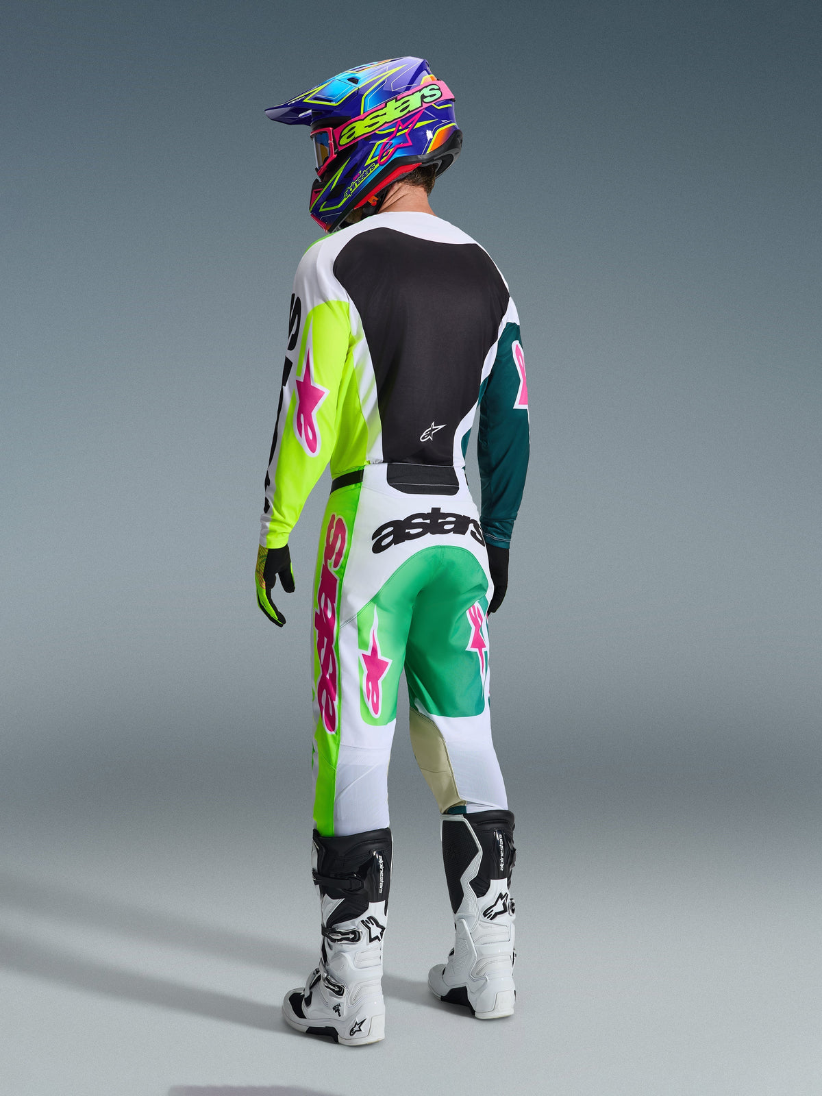 Alpinestars 2026 Racer Portl Jersey in Green White Black, motorcrosser van achteren gezien, bijpassende crossbroek met roze sterlogo's en 'astars' branding, witte en zwarte laarzen en een kleurrijke helm, complete gecoördineerde outfit voor off-road racing