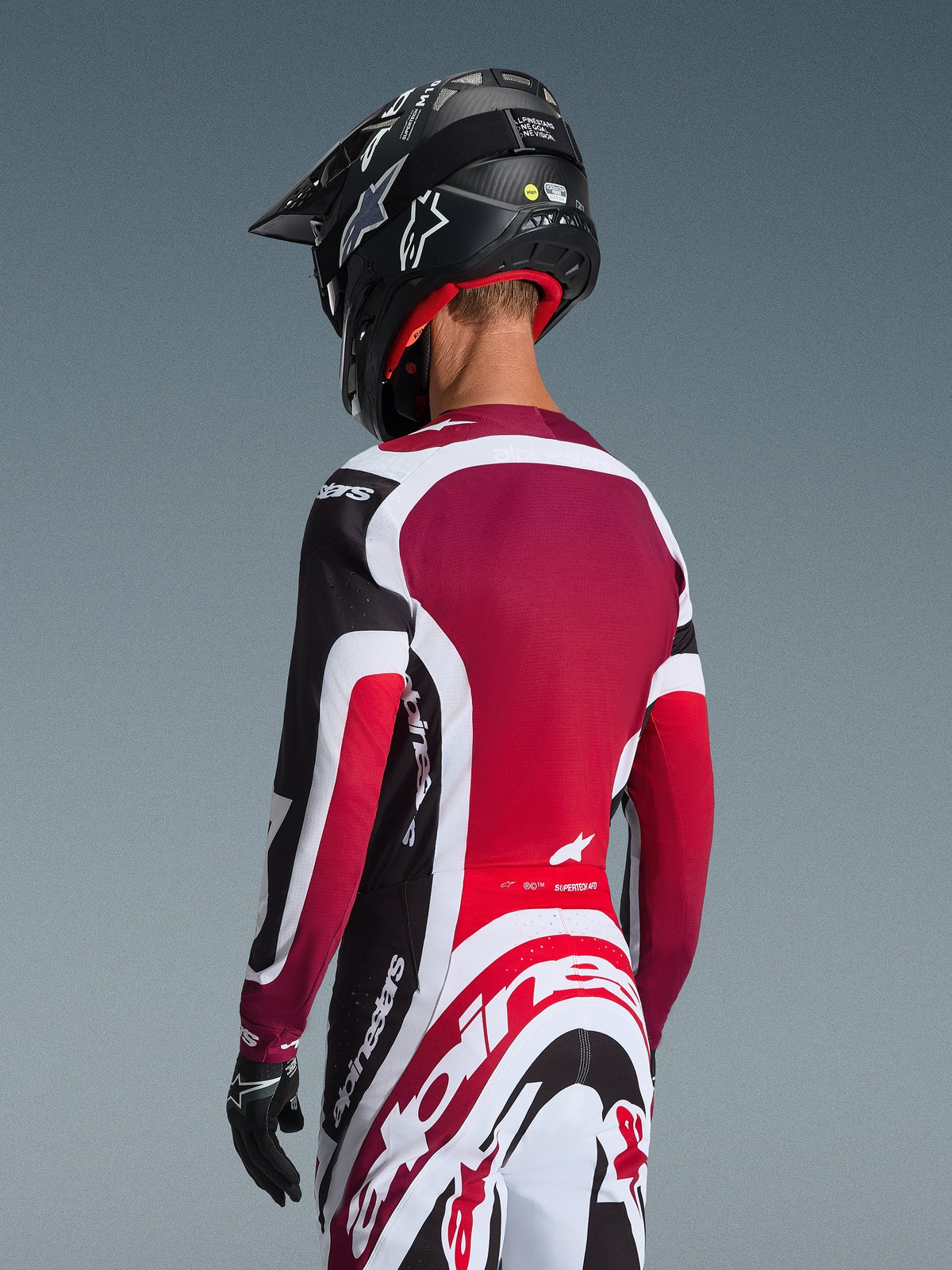 Alpinestars 2026 Supertech AFD Vista Gear Set, achteraanzicht van een motorcrosser in zwart, rood en witte racekleding, jersey met dieprood rugpaneel en atletische pasvorm, bijpassende broek met opvallend Alpinestars-logo, zwarte helm en handschoenen, professionele motorcross-outfit