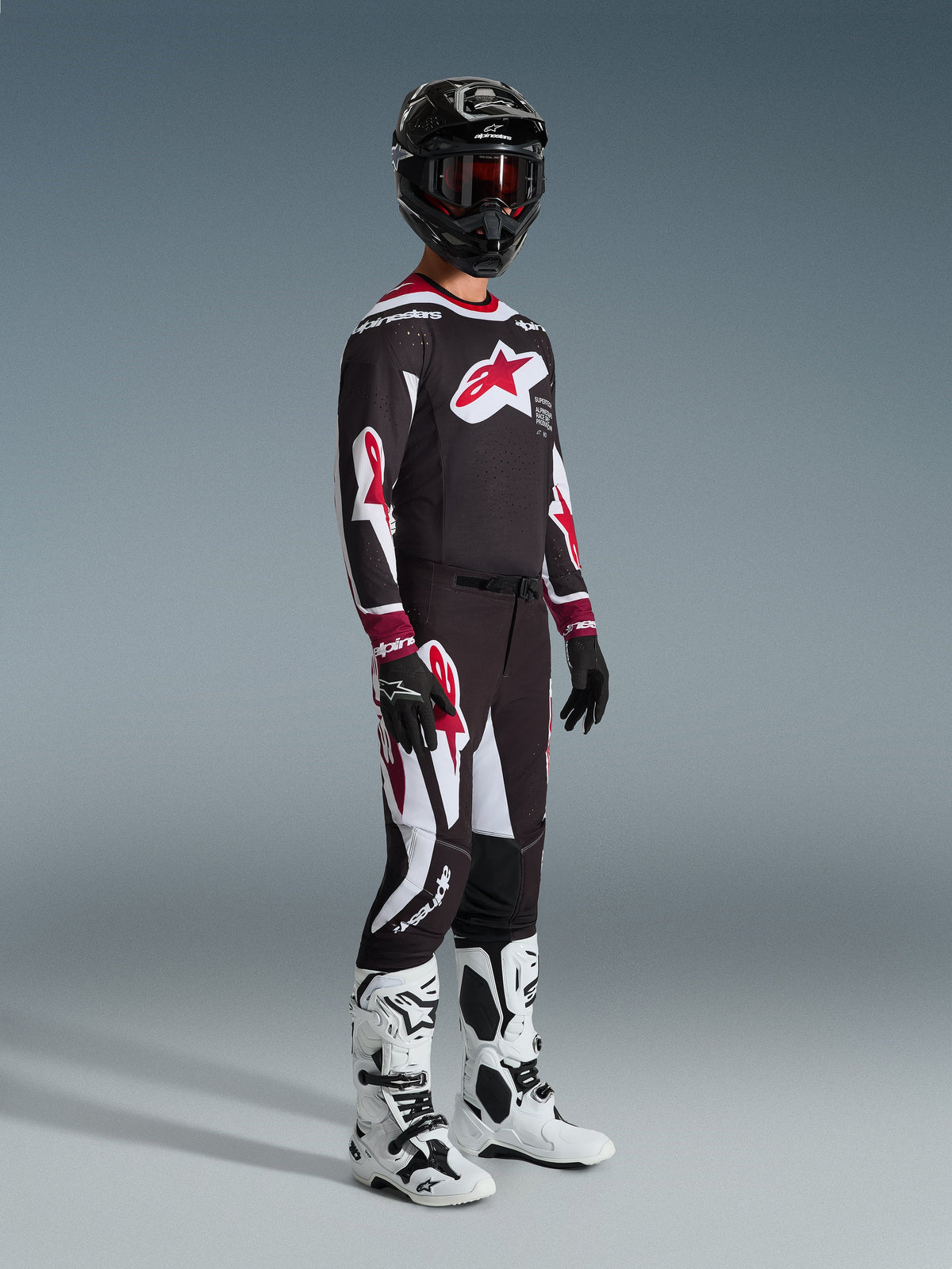 Alpinestars 2026 Supertech AFD Vista Gear Set, motorcrosser in volledige race-uitrusting, zwarte jersey met groot rood sterlogo op de borst, bijpassende zwarte broek met rode en witte details, witte en zwarte motorcrosslaarzen, zwarte helm met rode crossbril, professionele motorcross race-outfit