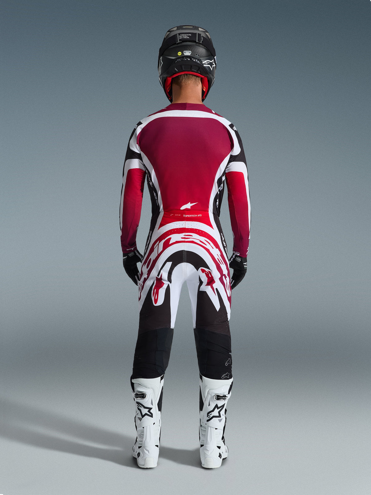 Alpinestars 2026 Supertech AFD Vista Gear Set, achteraanzicht van een motorcrosser in professionele race-uitrusting, bordeauxrood en rood shirt met witte en zwarte accenten, bijpassende broek met opvallende graphics, witte laarzen en zwarte helm, gecoördineerd motorcross racetenue.