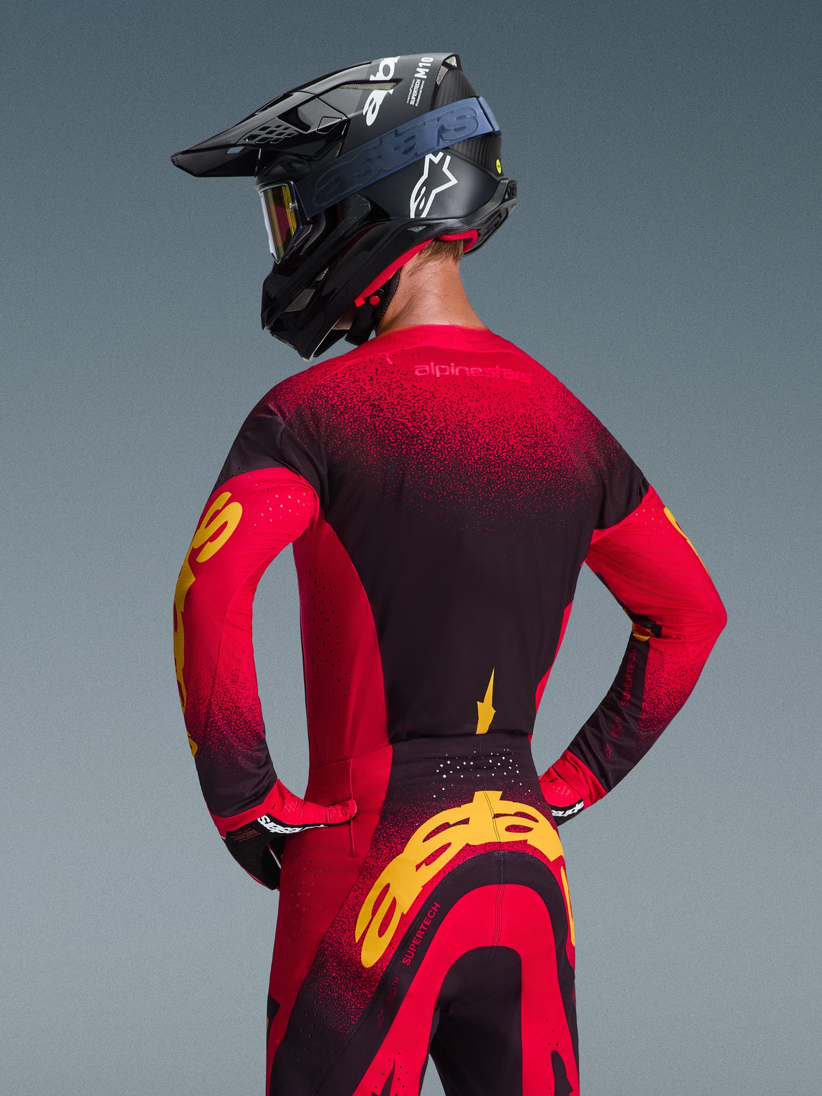Motocross rider viewed from the back wearing the Alpinestars 2026 Supertech Scenz Jersey and matching pants, Red Black and Yellow color scheme, black racing helmet with white logo, featuring bold yellow branding on the sleeves and rear, professional off-road gear.