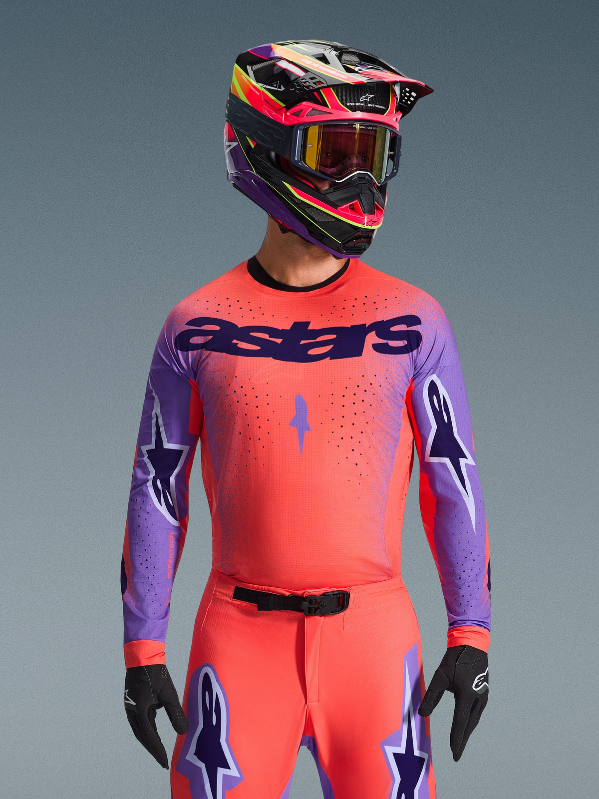 Alpinestars 2026 Supertech Scenz Jersey, motorcross race-jersey, Hot Coral en paars, voorzien van groot ‘astars’ logo en sterontwerpen op de mouwen, gedragen met bijpassende broek en een meerkleurige helm, hoogwaardige off-road uitrusting.