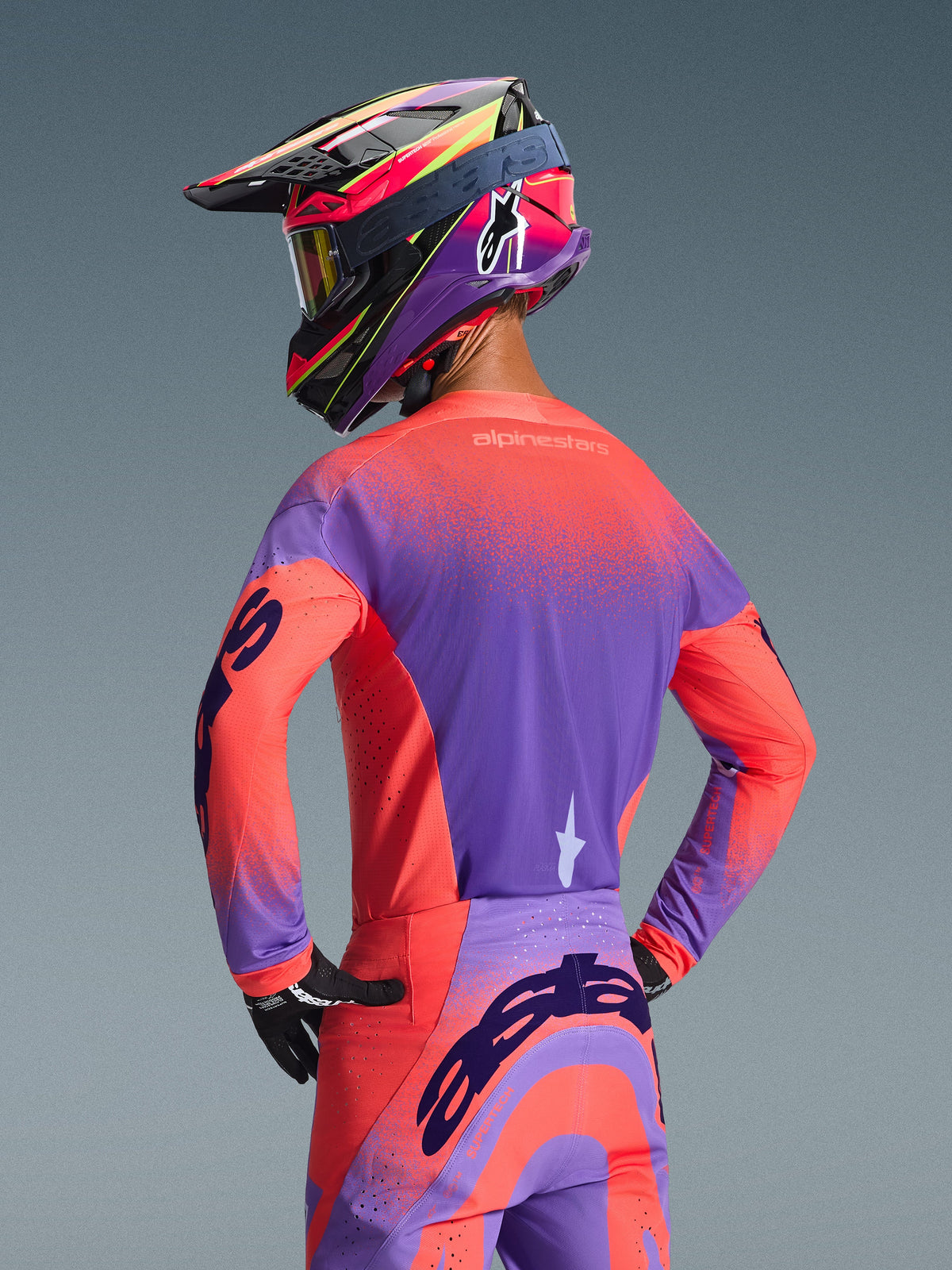 Motocrosser van achteren gezien in de 2026 Alpinestars Supertech Scenz Jersey in Hot Coral en Purple, voorzien van een opvallend gespikkeld kleurverloop, gecombineerd met een bijpassende motorcrossbroek en kleurrijke racehelm, professionele uitrusting voor off-road prestaties.
