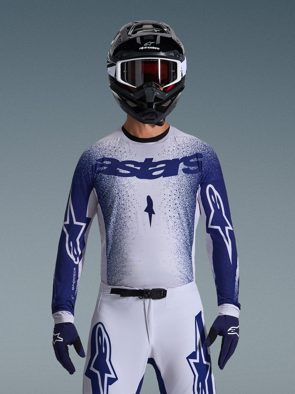 Alpinestars 2026 Supertech Scenz Gear Set, motocrossrijder in wit jersey met blauw gespikkeld kleurverloop en 'astars'-branding, bijpassende witte broek met blauwe accenten, zwarte helm met witte crossbril en blauwe handschoenen, atletische pro-level racepasvorm