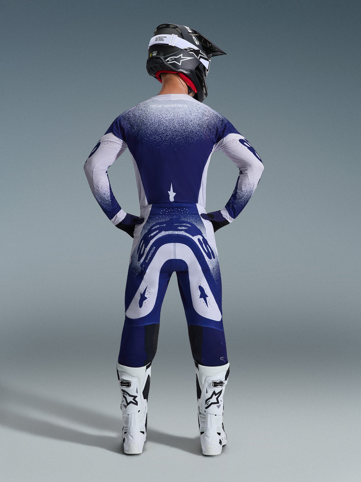 Alpinestars 2026 Supertech Scenz Gear Set, achteraanzicht van een motorcrosser in blauw-witte race-jersey en broek, gestippeld kleurverloop, gedragen met witte motorcrosslaarzen en een zwarte helm, professionele crossuitrusting
