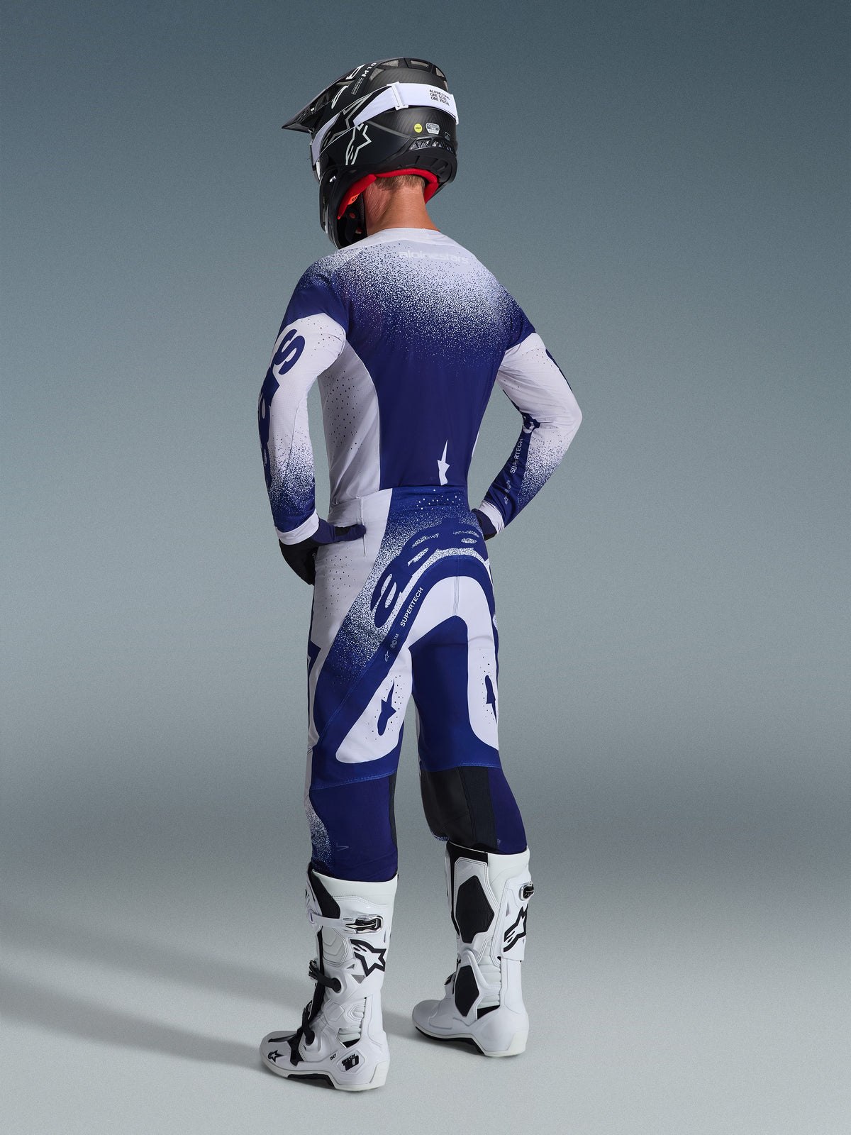 Motocrossrijder die de Alpinestars 2026 Supertech Scenz gearset draagt, marineblauwe en witte jersey en broek met een gespikkeld verloop-effect, achteraanzicht toont de atletische pasvorm, gecombineerd met witte en zwarte laarzen en een zwarte helm, professionele racekleding voor off-road prestaties
