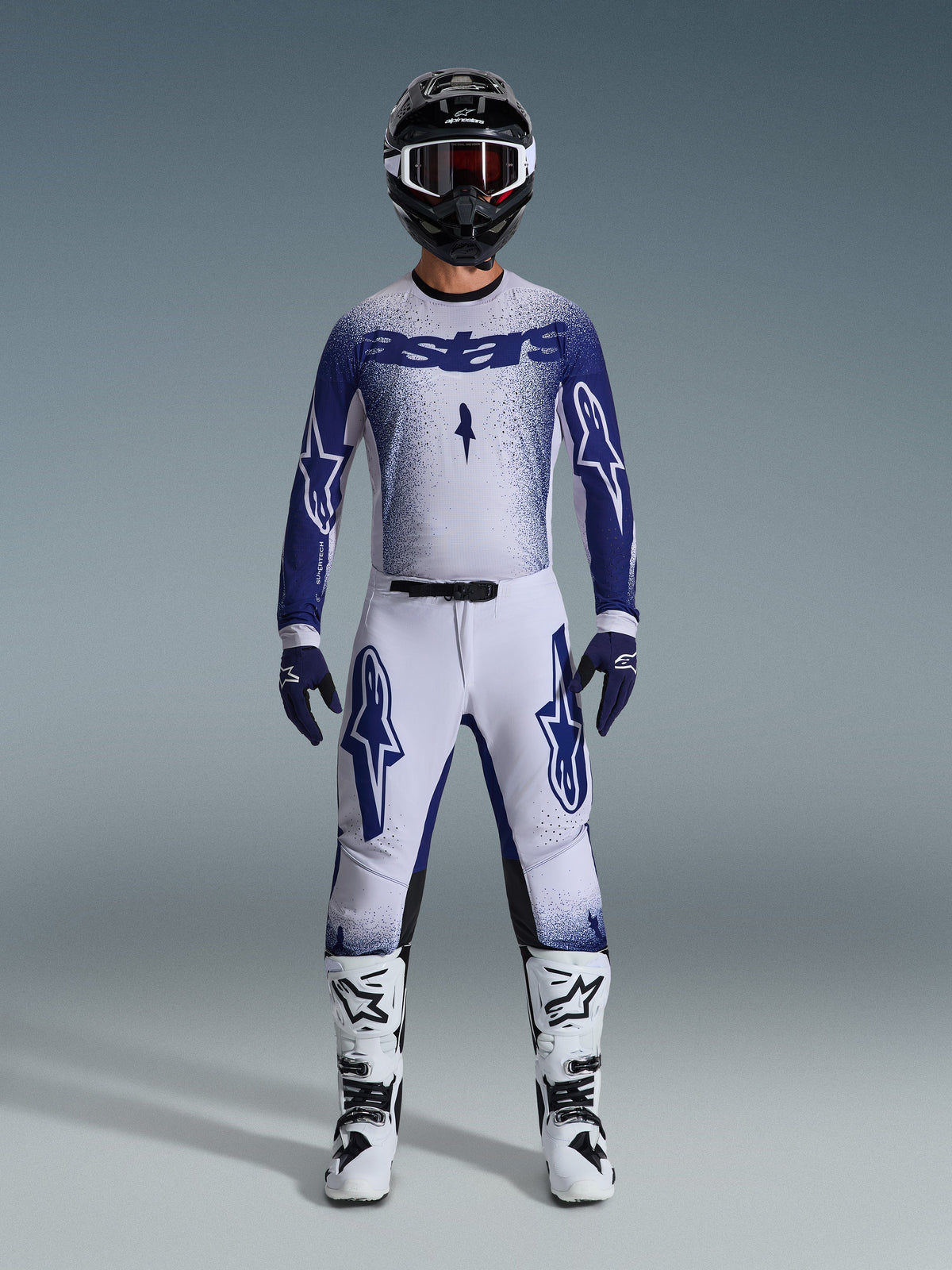 Motorcrosser in volledige race-uitrusting, 2026 Supertech Scenz Jersey in lichtgrijs en donkerblauw, bijpassende lichtgrijze broek met blauwe logo's, zwart-witte helm met rode crossbril, wit-zwarte laarzen, donkerblauwe handschoenen, gecoördineerde outfit voor off-road racen