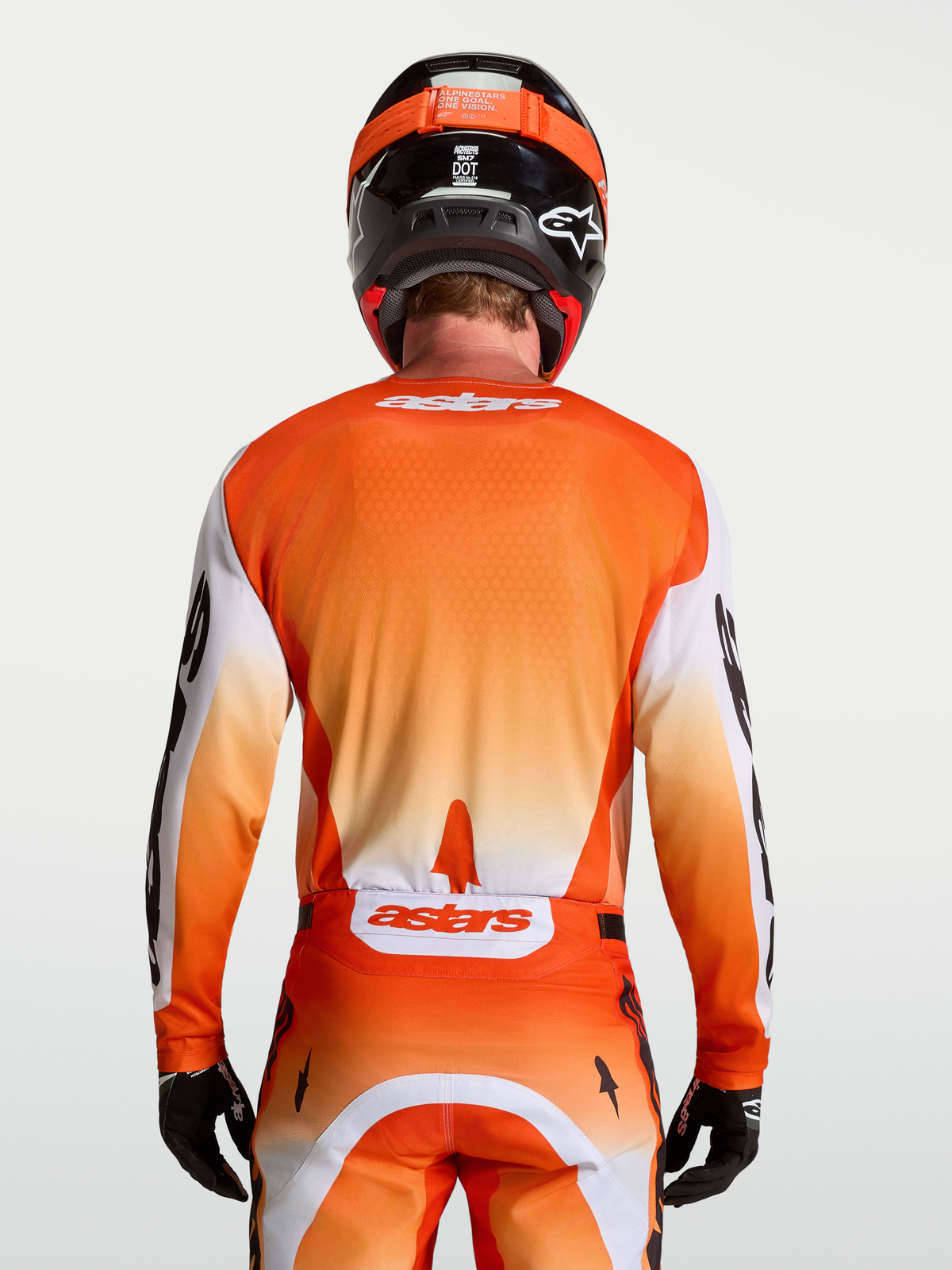 Alpinestars Racer Amber jersey, ademende motocross jersey voor heren, oranje en wit, achteraanzicht van rijder in volledige off-road race-uitrusting, vochtafvoerend technisch materiaal.