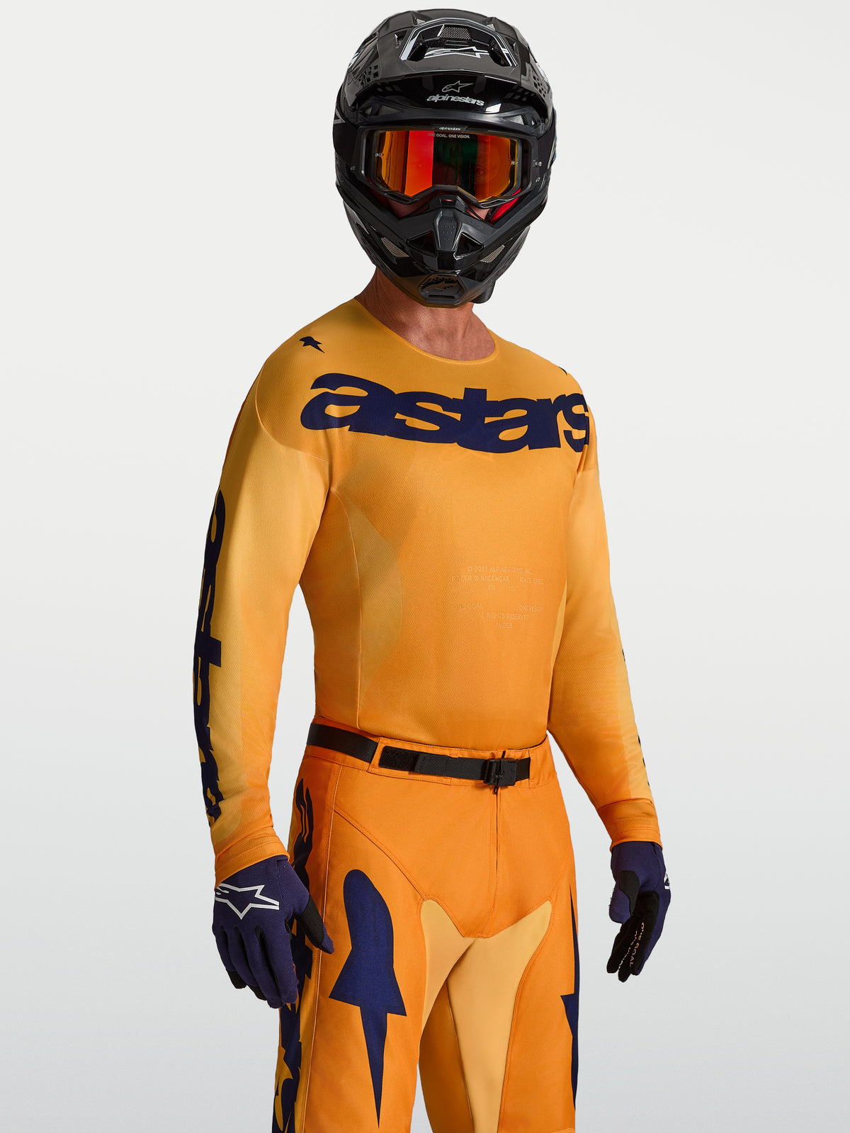 Alpinestars Racer Amber Jersey, vochtafvoerende motocrossjersey voor heren in oranje goud en diepblauw, gedragen door een model met MX-helm en handschoenen.
