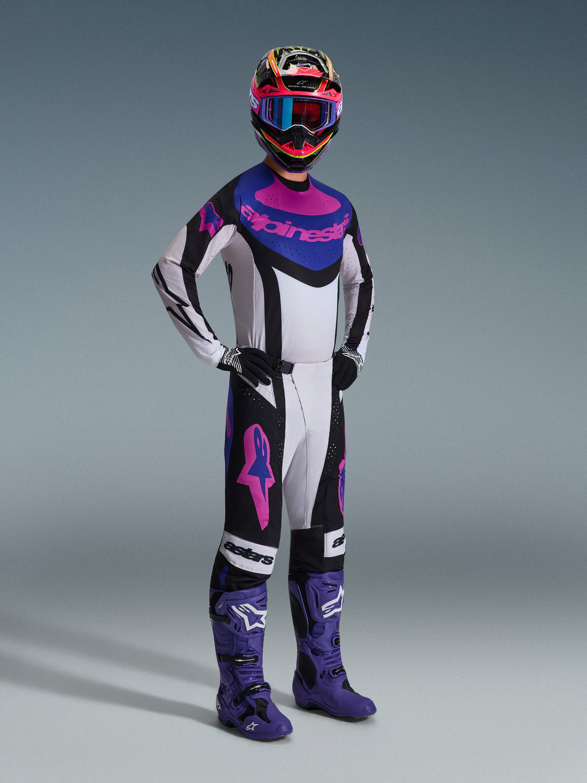 Motocross rider wearing the Alpinestars 2026 Techstar Knif Jersey in Black Light Gray Purple, matching pants with large pink logos, purple boots, and a multi-colored helmet with blue goggles, full racing kit for off-road performance