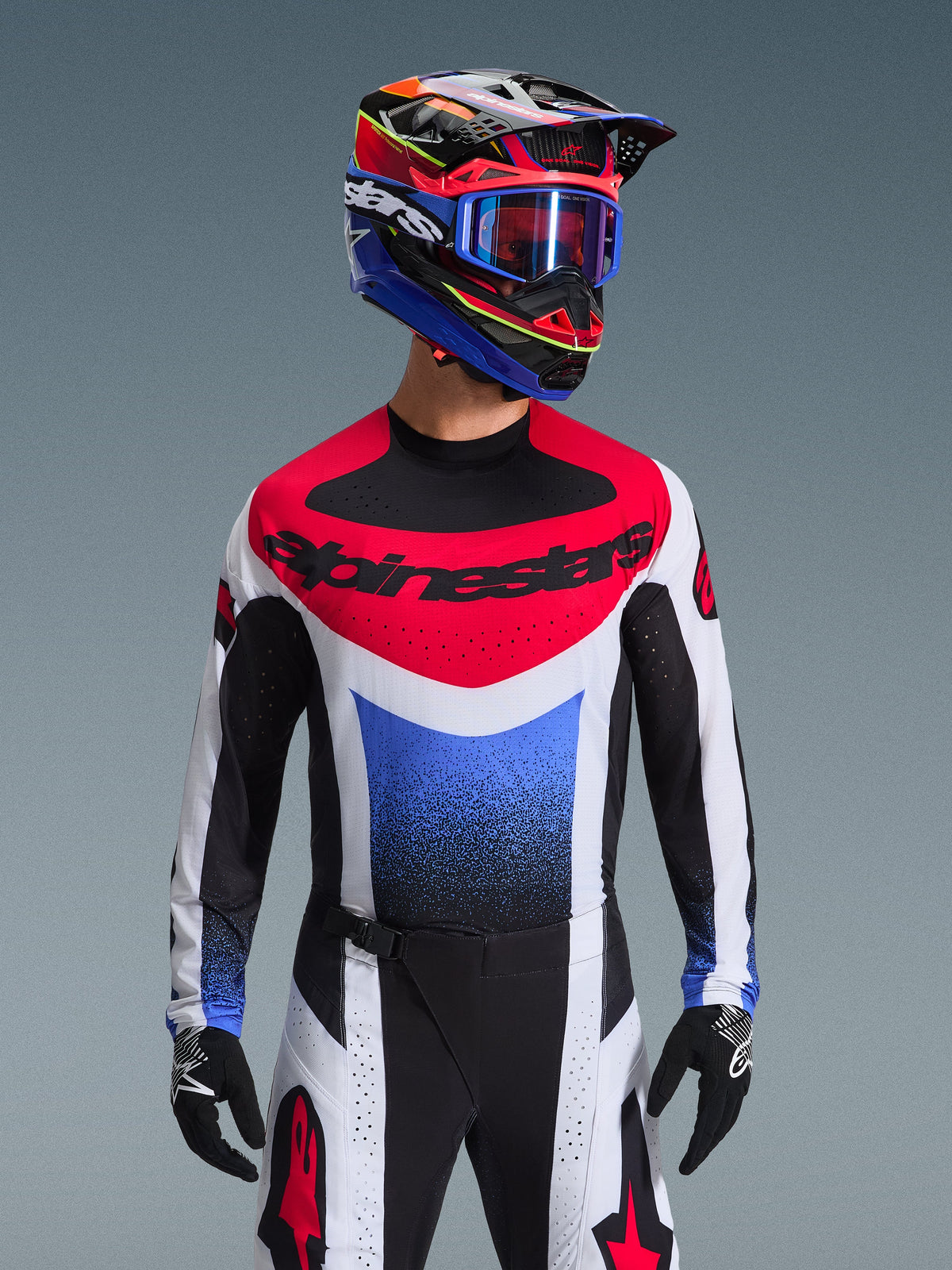 Alpinestars 2026 Techstar Knif Gear Set, motorcrosser met volledige race-uitrusting, rood-wit-blauw shirt met grote zwarte branding, bijpassende zwart-witte broek met rood sterlogo, meerkleurige helm met blauwe crossbril en zwarte handschoenen, gecoördineerde outfit voor off-road competitie