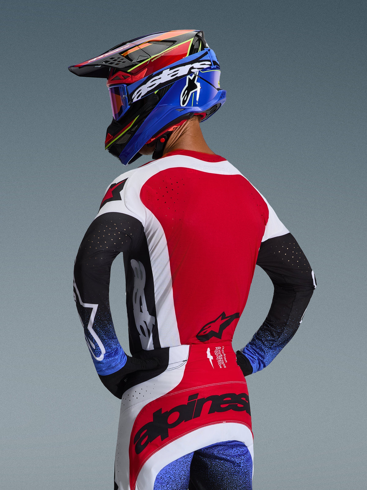Motocrossrijder van achteren gezien in de 2026 Techstar Knif Jersey in rood, wit en zwart, met lasergesneden perforaties op de rug en opvallende branding, gecombineerd met een bijpassende broek met blauwe accenten en een blauw-zwarte Alpinestars helm, professionele off-road race-uitstraling