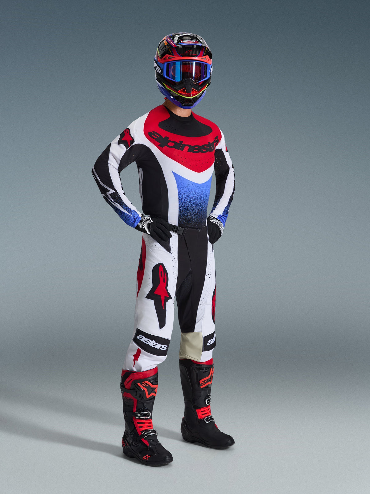 Motocrosser in de Alpinestars 2026 Techstar Knif Gear Set, jersey met zwarte, witte en rode panelen en een blauw kleurverloop op de borst, witte broek met rode Alpinestars-logo's en zwarte accenten, zwarte en rode laarzen, complete professionele off-road race-uitrusting.