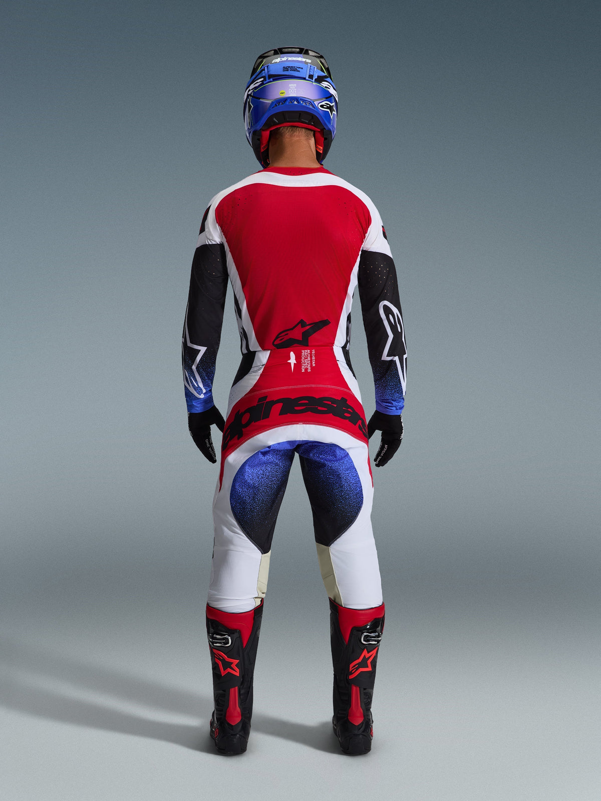 Alpinestars 2026 Techstar Knif Gear Set, complete motorcross outfit van achteren gezien, rood-wit-zwart jersey met blauwe kleurverloopaccenten op de mouwen, bijpassende witte en zwarte broek met opvallende rode branding, blauwe helm en zwarte en rode laarzen, professionele off-road racing outfit