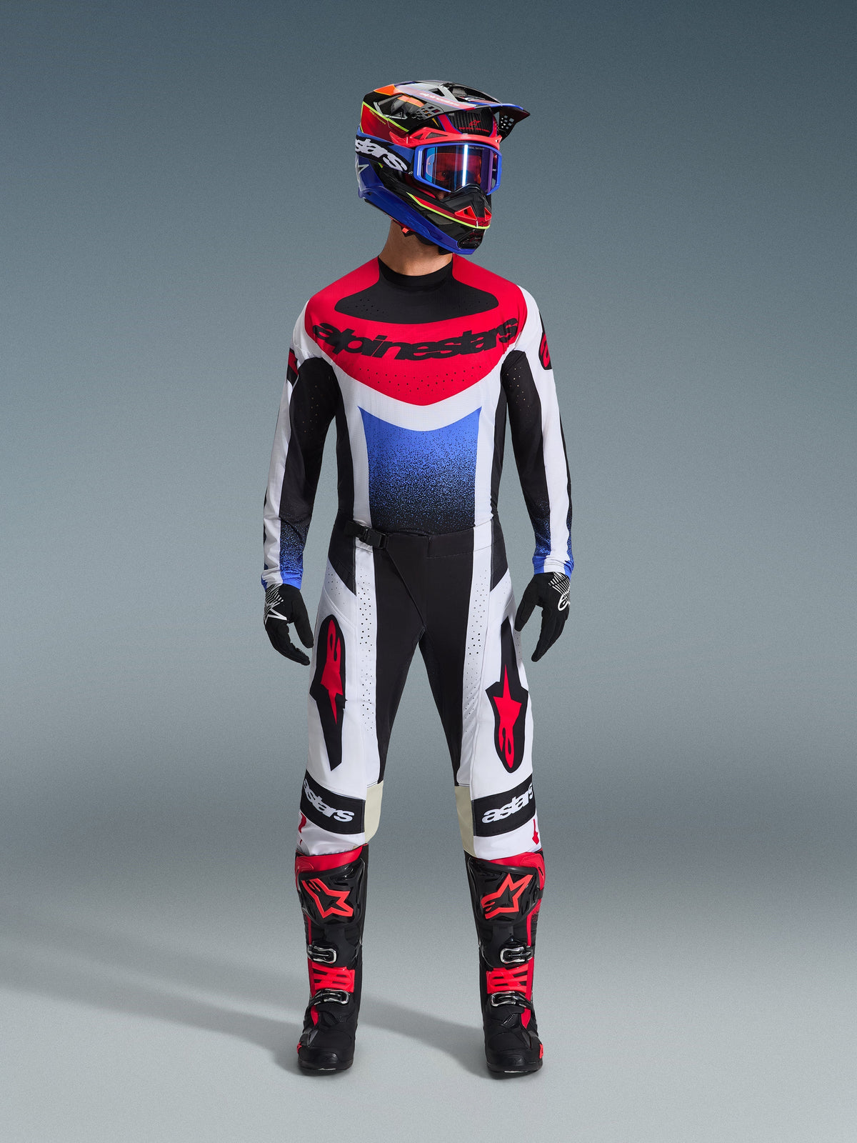 Motocrossrijder in de Alpinestars 2026 Techstar Knif Gear Set, zwart, wit en rood shirt met blauw kleurverloop en branding, bijpassende zwart-witte broek met rode accenten, meerkleurige helm met blauwe crossbril en zwarte en rode laarzen, gecoördineerde off-road race-outfit.
