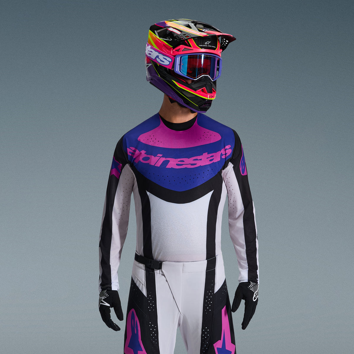 Alpinestars 2026 Techstar Knif Gear Set, motocross-rijder met jersey met blauwe en roze borstgrafiek en wit torso, witte broek met zwarte panelen en roze logo's, veelkleurige helm in paars en roze met bijpassende crossbril, zwarte handschoenen, professionele off-road race-uitrusting