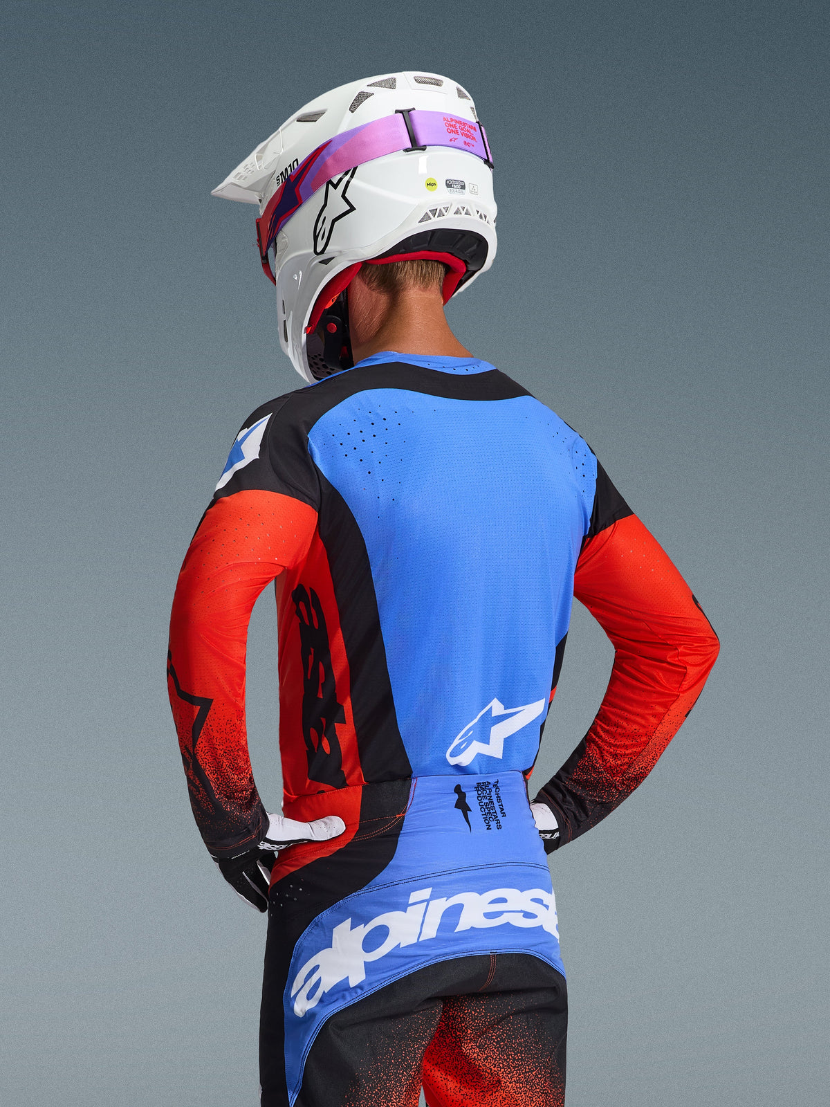 Motorcrosser van achteren gezien in de 2026 Techstar Knif Gear Set, blauwe jersey met rode mouwen en zwarte accenten, zwarte en blauwe broek met witte Alpinestars-branding, witte helm met roze brilband, voorzien van lasergesneden perforaties voor ventilatie
