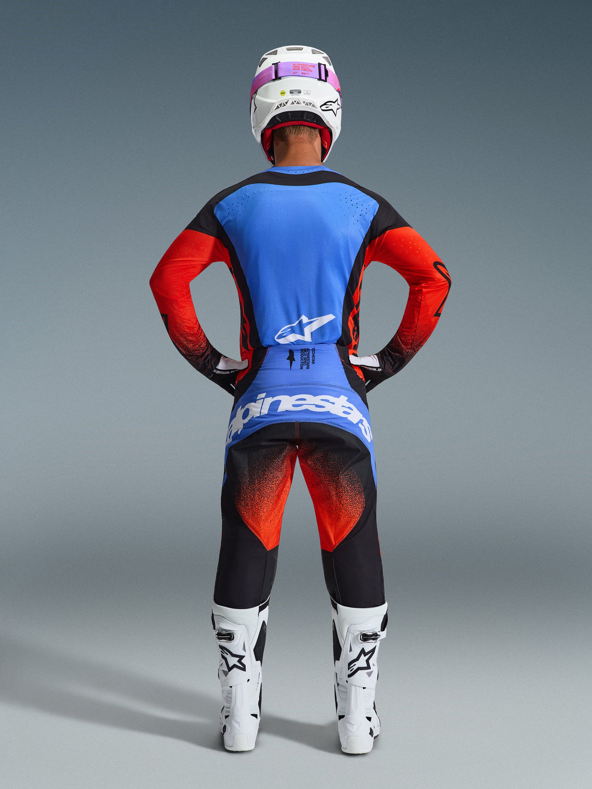 Achteraanzicht van een motorcrosser in de Alpinestars 2026 Techstar Knif Gear Set, blauwe en rode jersey met zwarte accenten, zwarte broek met rood kleurverloop op de dijbenen en blauwe branding, gecombineerd met witte racelaarzen en een witte helm met roze bril.