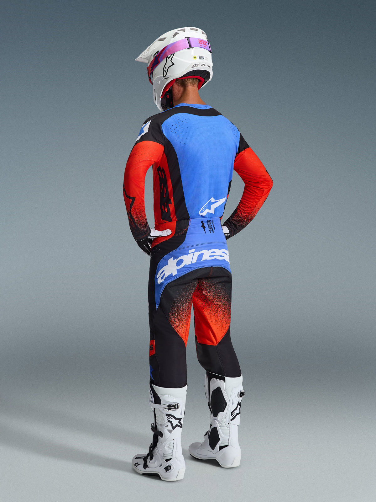 Alpinestars 2026 Techstar Knif Gear Set, achteraanzicht van een motorcrosser, blauwe en rode jersey met zwarte accenten, bijpassende zwarte en rode broek met een groot blauw logo op het zitvlak, witte helm en witte motorcrosslaarzen, professionele off-road race-uitrusting