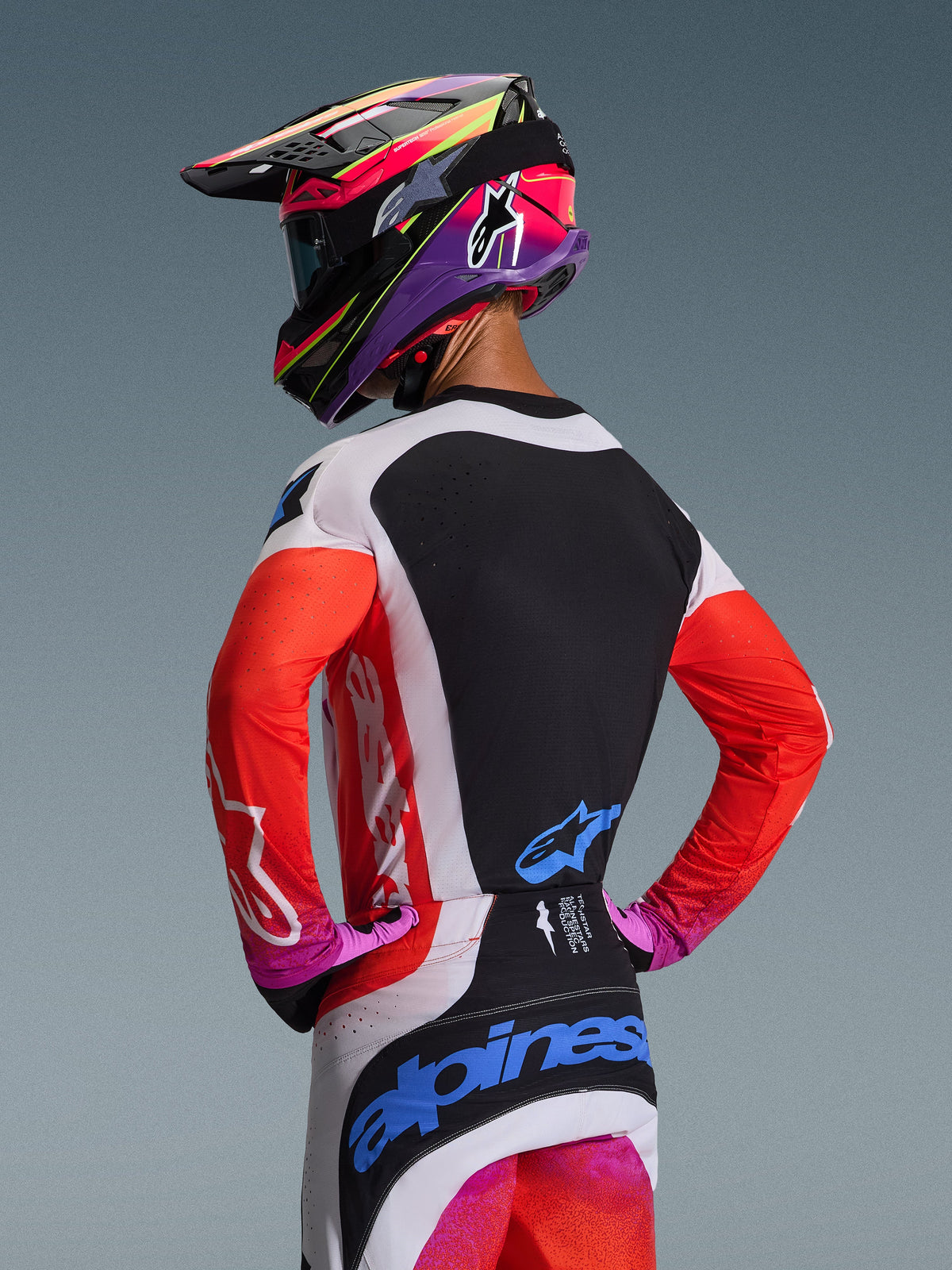 Motorcrossrijder van achteren in de Alpinestars 2026 Techstar Knif Jersey in Light Gray Orange Fluo Black, met een zwart rugpaneel met lasergesneden ventilatie, oranje mouwen en lichtgrijze zijpanelen, gecombineerd met een bijpassende broek en een kleurrijke helm.
