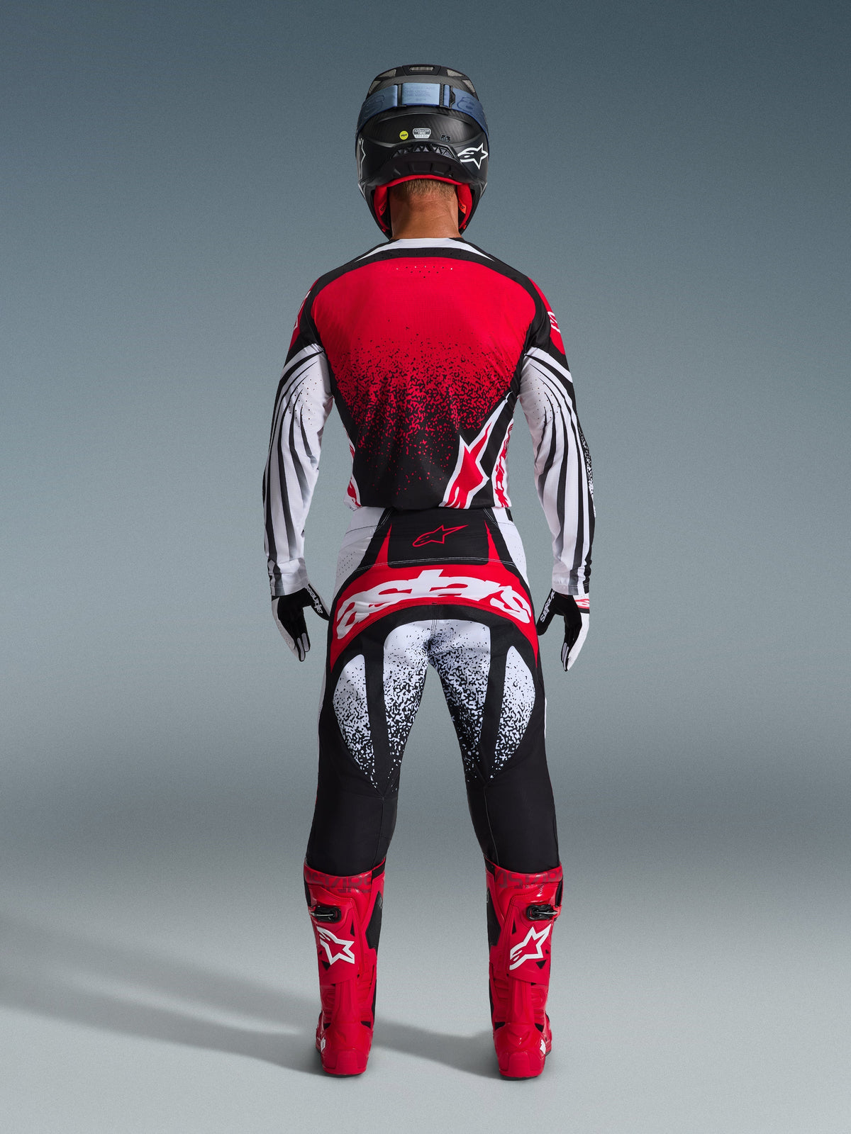 Motocross rider from behind wearing the Alpinestars 2026 Techstar Nomur Jersey, Red Black and White with stippled pattern, matching racing pants, red motocross boots and black helmet, complete gear set for off-road competition