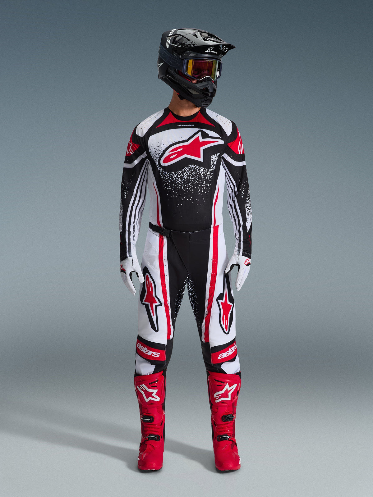 Motocross rider wearing full Alpinestars racing gear, 2026 Techstar Nomur Jersey in Black White Red, matching pants with red accents, black helmet, and bright red boots, features large red Astars logo on chest with speckled gradient pattern, coordinated outfit for off-road competition