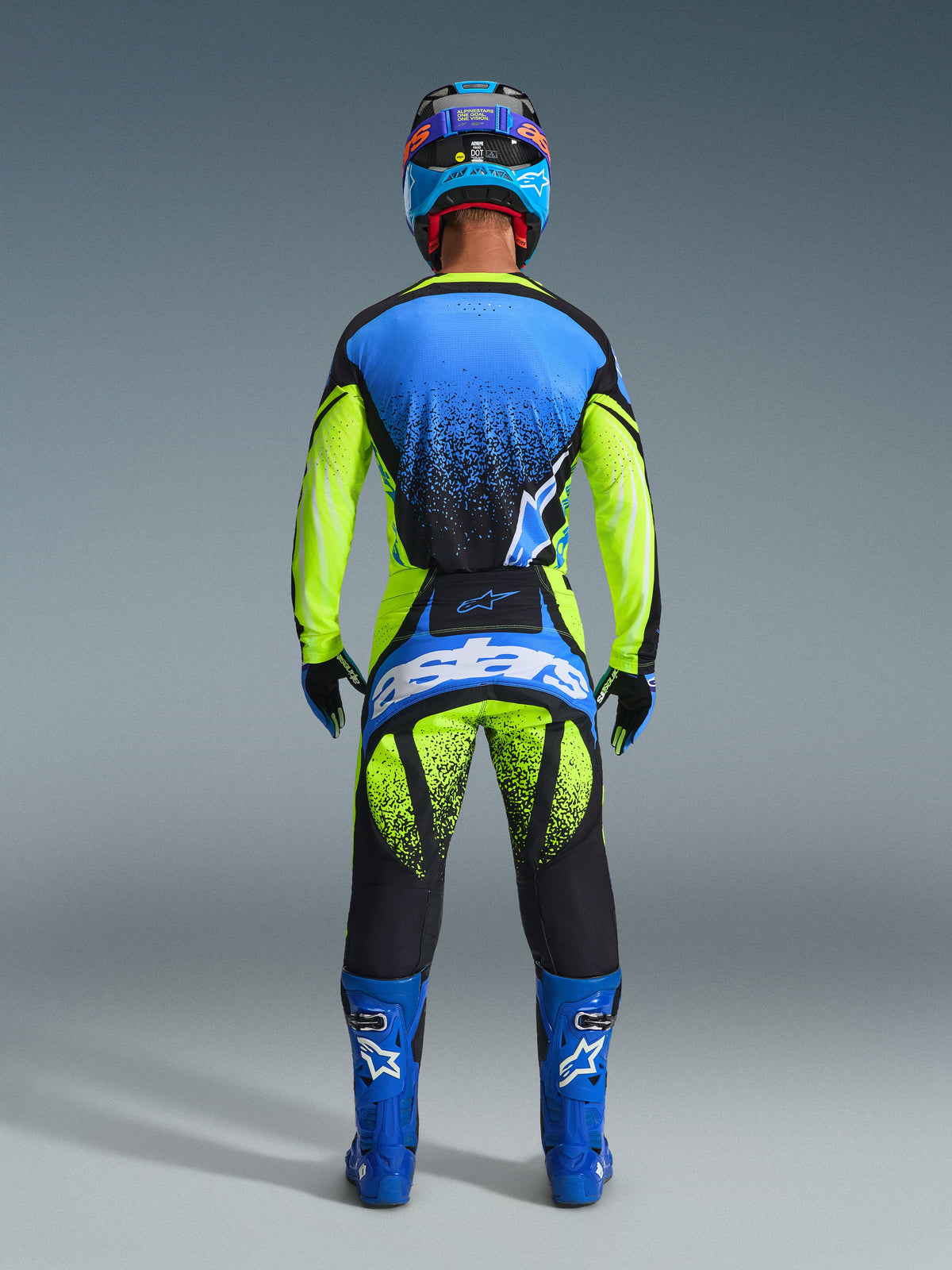 Rear view of a motocross rider wearing the Alpinestars 2026 Techstar Nomur Gear Set. The outfit includes a blue and neon yellow jersey with black spray-pattern accents, matching neon yellow and black pants featuring a large blue 'astars' logo on the seat, coordinated blue boots with white logos, and a blue and black helmet. Full professional racing gear designed for performance and visibility.