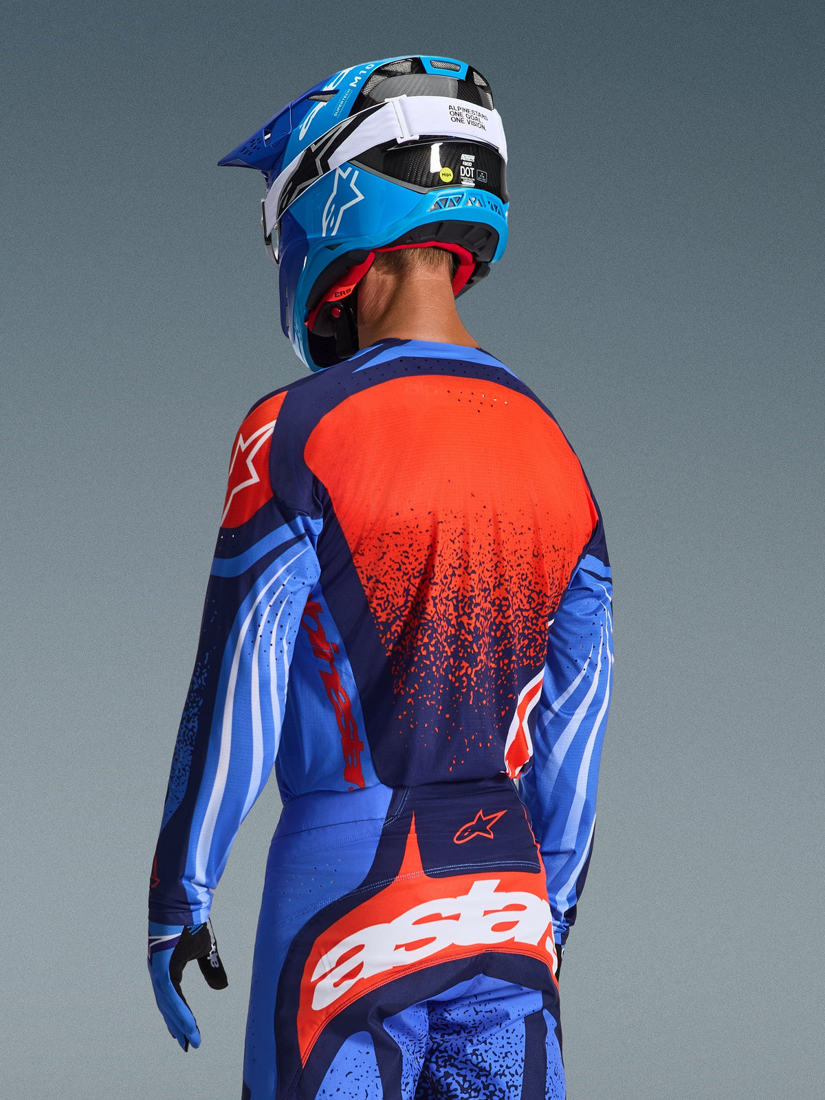 Alpinestars Techstar Nomur Jersey 2026, motorcrossrijder van achteren gezien in donkerblauw met oranje en UCLA-blauwe accenten, voorzien van een grote oranje grafische print met textuur op de rug, gecombineerd met een bijpassende motorcrossbroek en een blauwe racehelm, professionele uitrusting voor off-road rijden.