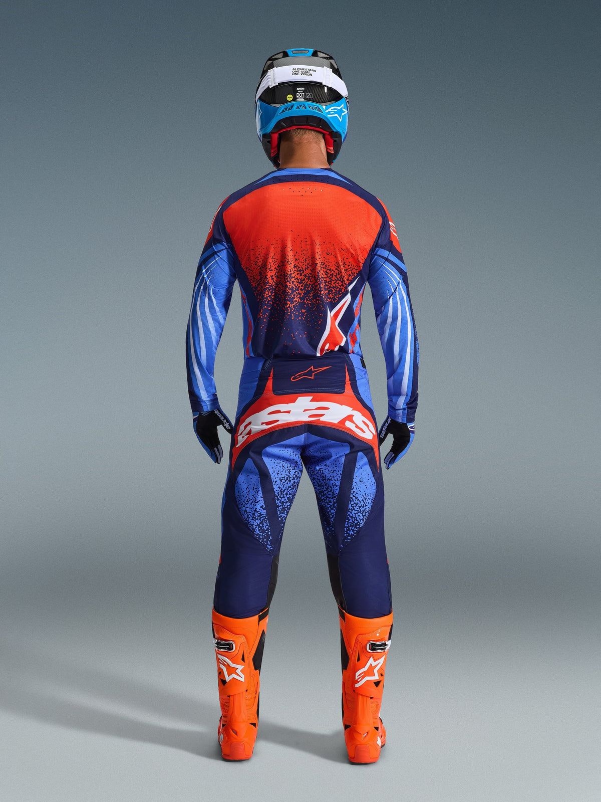Motocrosser van achteren gezien in de 2026 Techstar Nomur Jersey en bijpassende broek, kleurstelling in Dark Navy en Orange met UCLA Blue accenten, feloranje laarzen, blauw-witte helm en zwarte handschoenen, complete professionele off-road race-uitrusting