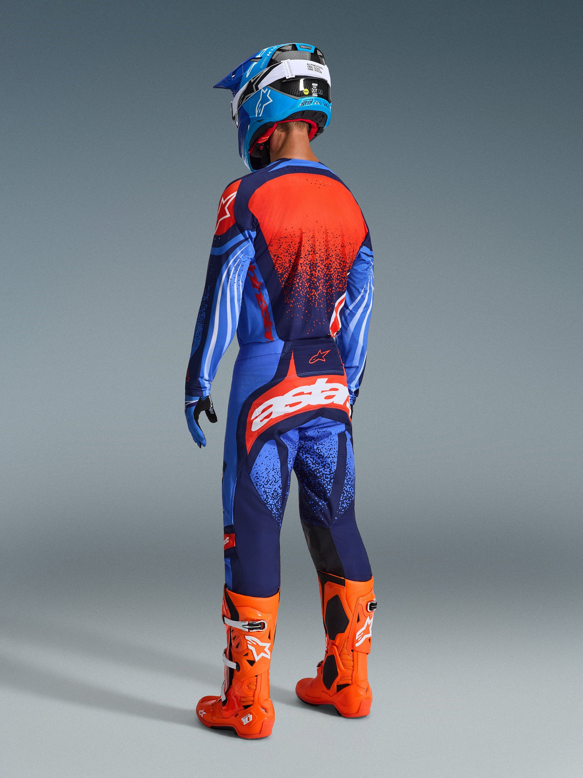 Motorcrossrijder van achteren gezien in de Alpinestars 2026 Techstar Nomur Jersey in Dark Navy, Orange en UCLA Blue, gecombineerd met een bijpassende broek met opvallende witte branding, feloranje laarzen en een blauw-witte helm, complete off-road race-uitrusting.