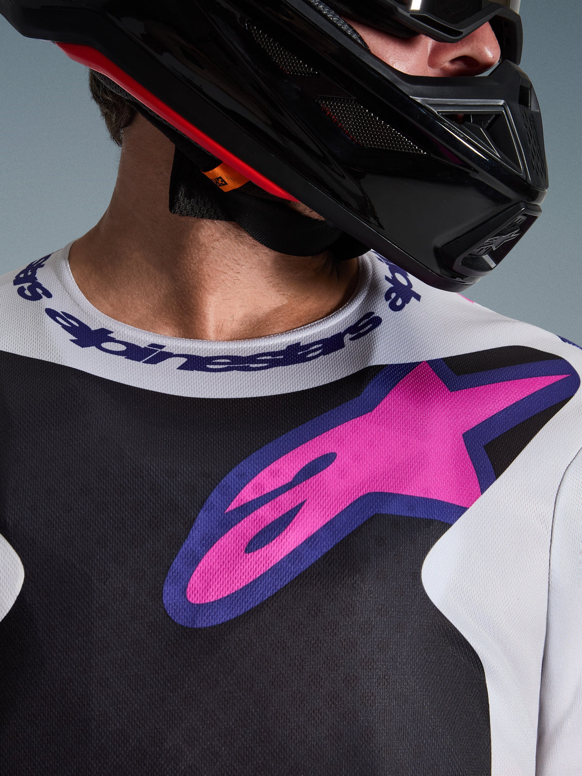 Close-up van de Alpinestars 2026 Fluid Grid Jersey, motorcross jersey, zwart, lichtgrijs en paars, met een opvallend roze-paars sterlogo op de borst en sportkraag met merknaam, gedragen met een zwarte motorcrosshelm