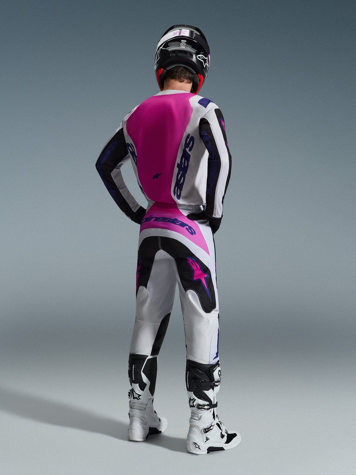 Alpinestars 2026 Fluid Grid Gear Set, motorcrosser van achteren gezien, witte jersey met een roze rugpaneel en paarse branding, bijpassende witte en zwarte broek met roze accenten, witte en zwarte laarzen en een zwarte helm, gecoördineerde off-road race-outfit.