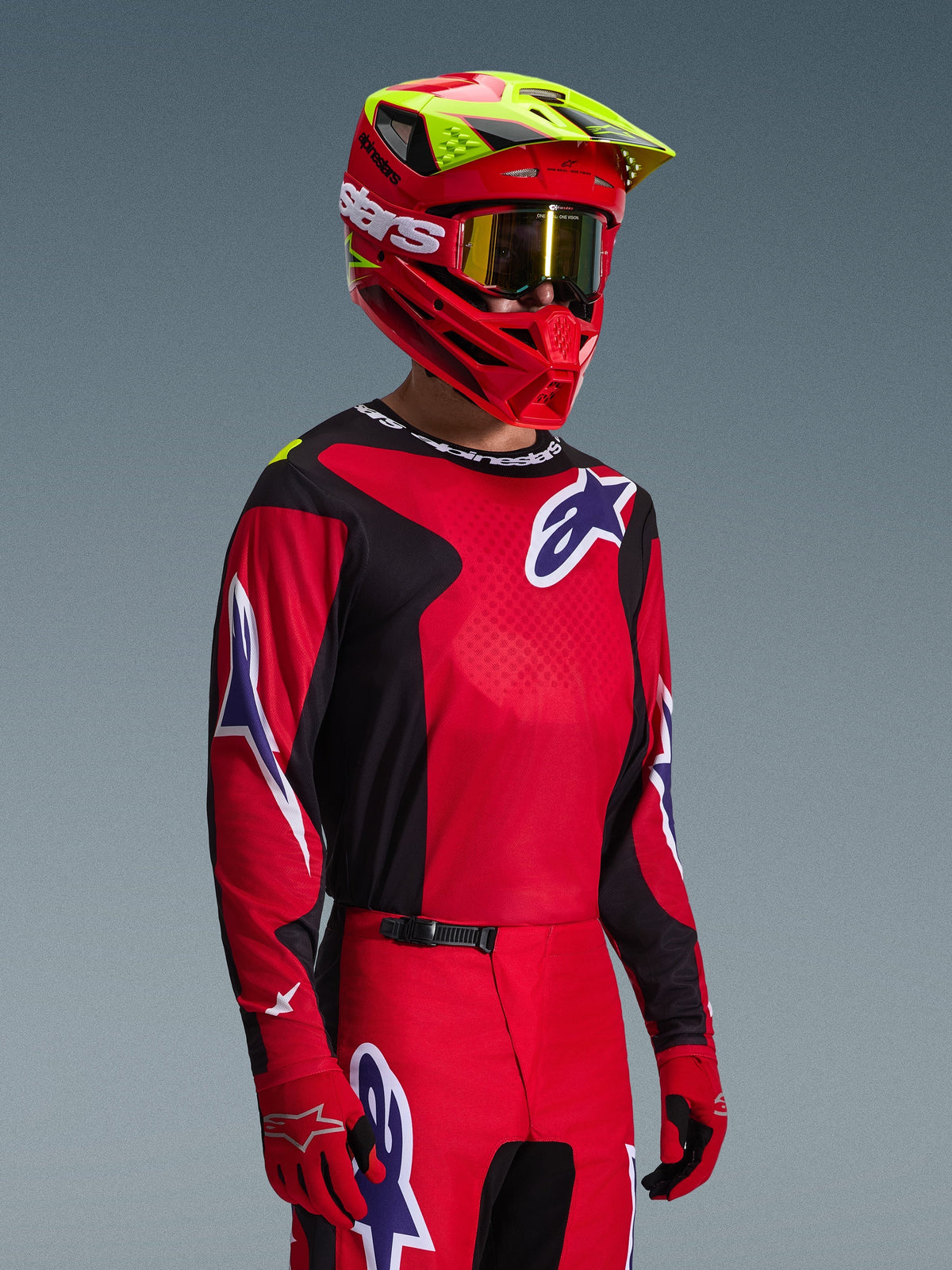 Motorcrosser in Alpinestars 2026 Fluid Grid Gear Set, rood en zwart shirt met witte en blauwe logo-accenten, bijpassende rood-zwarte broek, rode helm met gele accenten en gouden crossbril, gecoördineerde off-road race-uitrusting