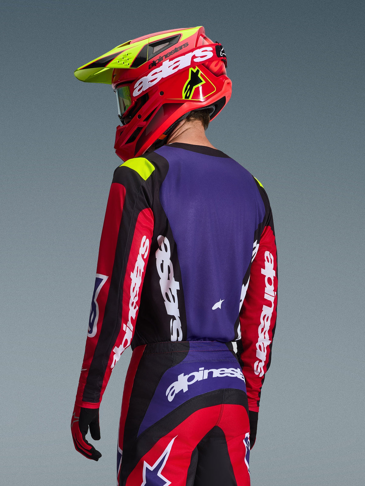 Motocrosser draagt Alpinestars 2026 Fluid Grid Jersey, rood, zwart en paars, achteraanzicht met paars rugpaneel en rode mouwen met witte branding, bijpassende broek en een rode en neongele helm, gecoördineerde outfit voor off-road racing
