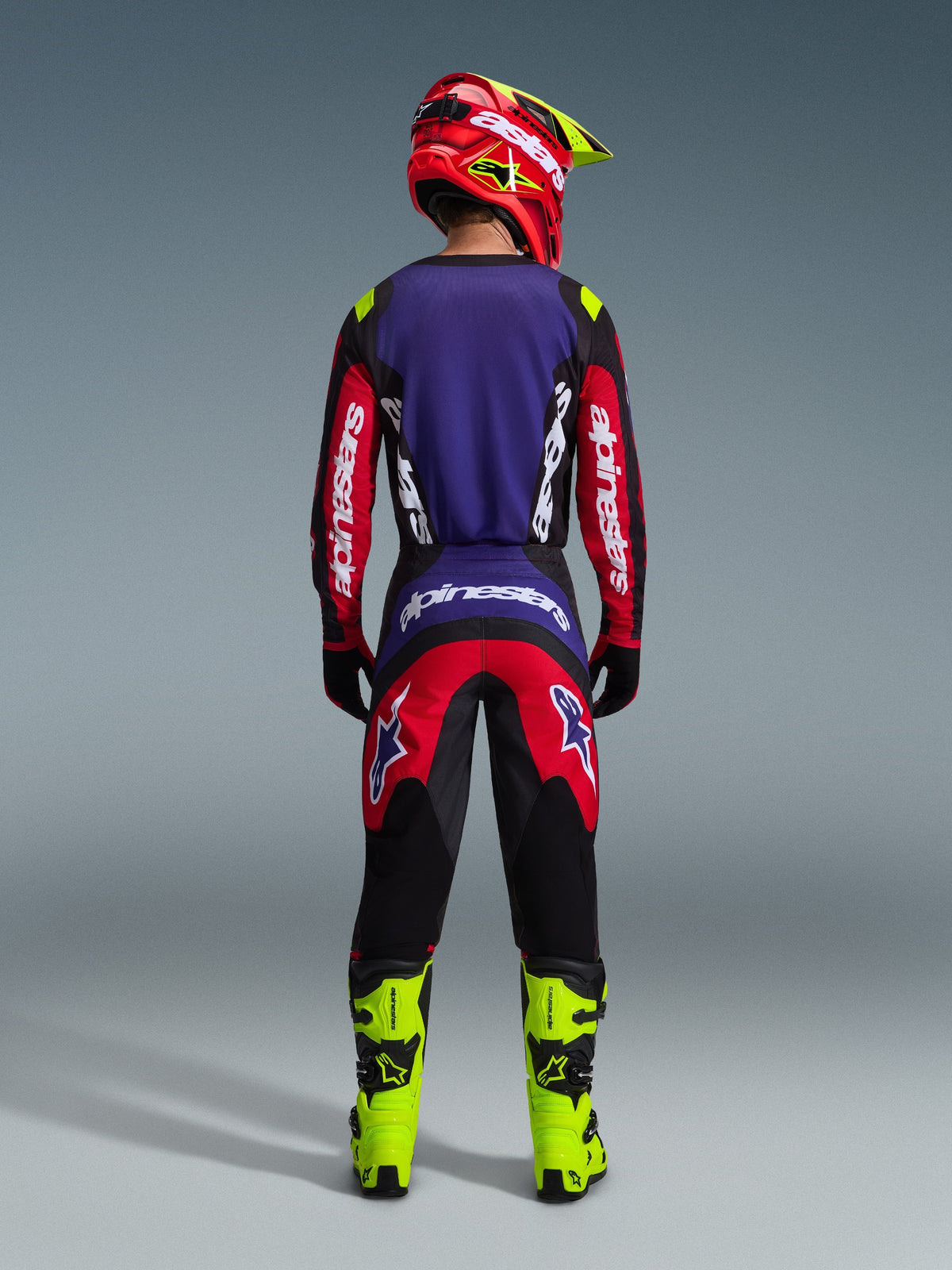 Motorcrosser van achteren gezien in de 2026 Fluid Grid Jersey in rood, zwart en paars, met witte Alpinestars-branding op de lange mouwen, gecombineerd met een bijpassende crossbroek, neongele laarzen en een rood-gele helm, complete uitrusting voor off-road racen