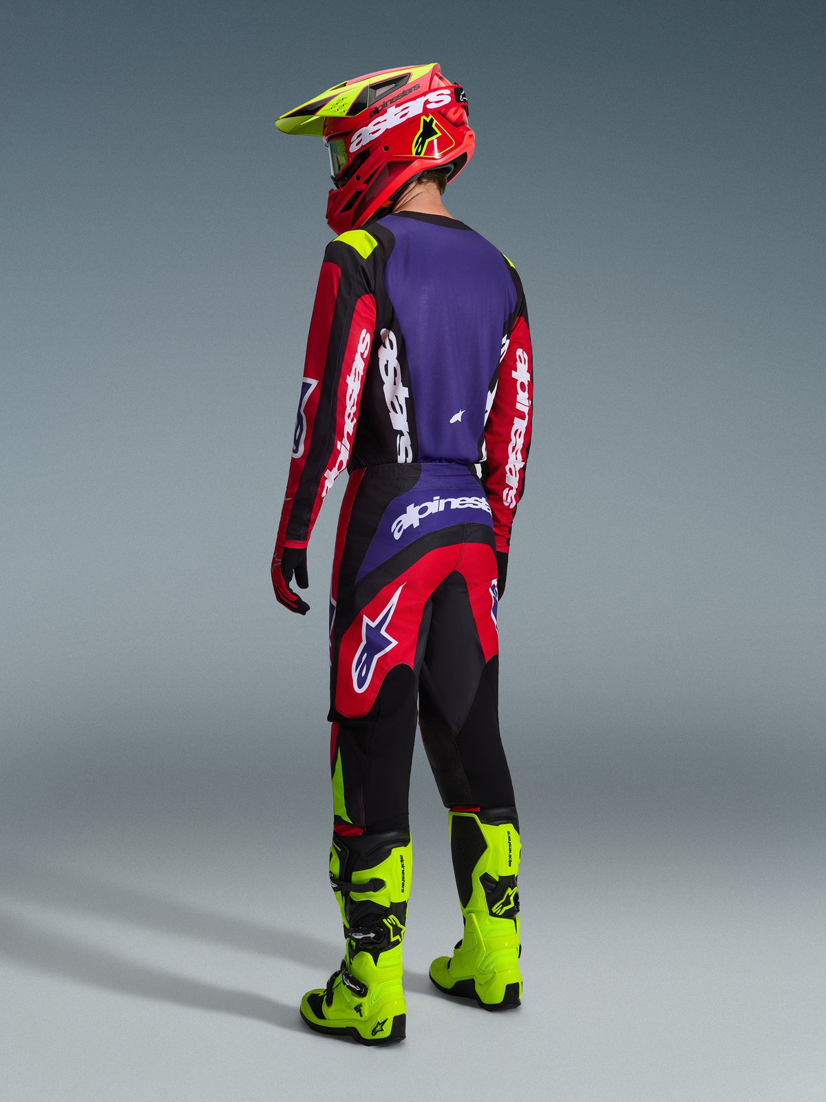 Alpinestars 2026 Fluid Grid Gear Set, motorcrosser van achteren gezien, rood, zwart en paars jersey met witte logo's, rode en zwarte broek met paarse accenten, gecombineerd met een rode en neongele helm en fel neongele laarzen, volledige off-road race-uitrusting