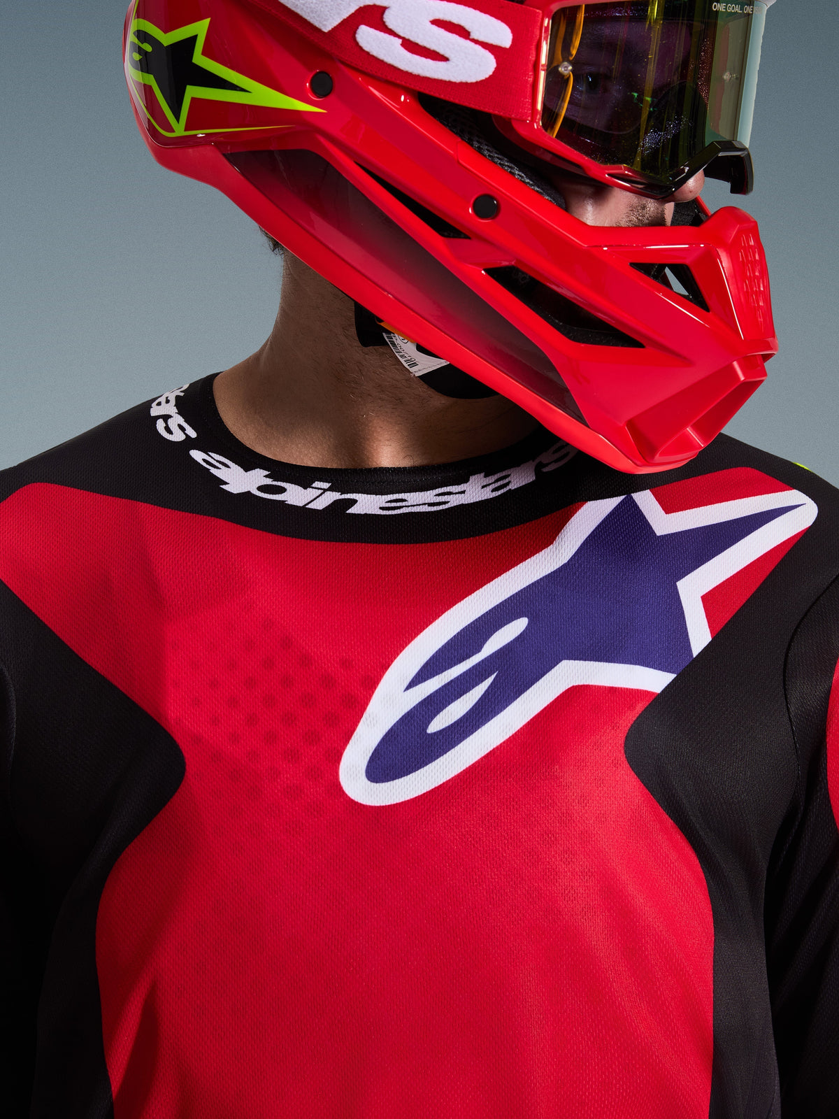 Close-up van een motorcrosser die de 2026 Fluid Grid Jersey in rood, zwart en paars draagt, met een groot Alpinestars-logo op de borst en een sportkraag met merknaam, gecombineerd met een rode helm en crossbril, wat de technische polyfabric en de atletische pasvorm benadrukt.