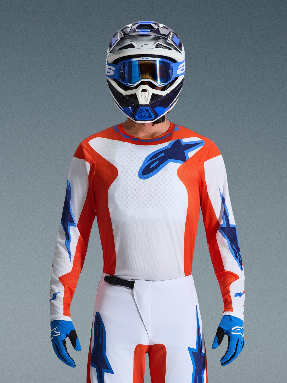 Alpinestars 2026 Fluid Grid Gear Set, motocrossrijder in oranje en wit jersey met blauwe ster-logo's, witte broek met blauwe accenten, bijpassende blauw-witte helm met blauwe crossbril en handschoenen, complete off-road race-outfit