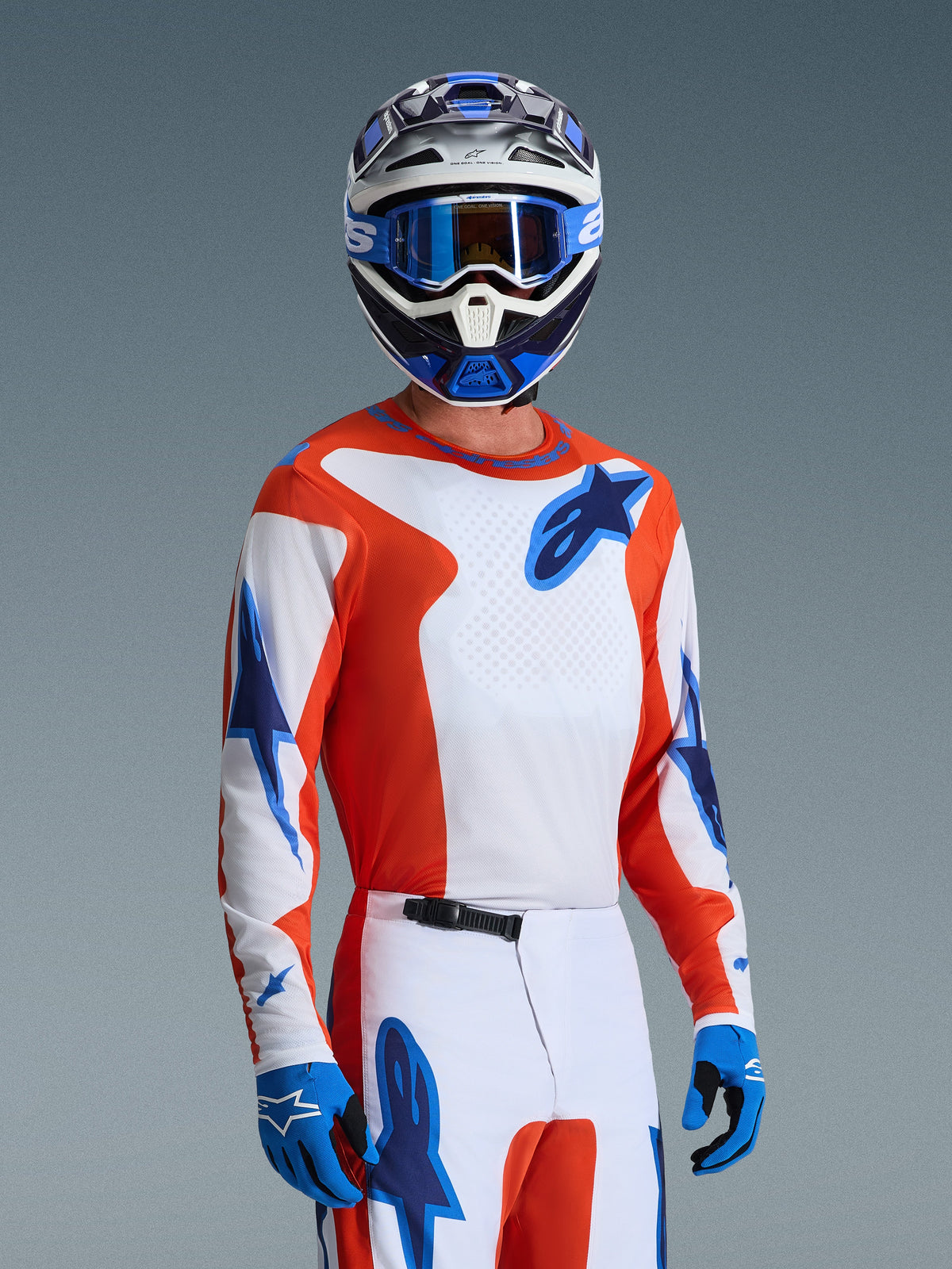 Motorcrosser met de Alpinestars 2026 Fluid Grid Gear Set, oranje en wit jersey met blauwe sterlogo's, witte en oranje broek met blauwe accenten, blauwe handschoenen, blauw-witte helm met blauwe crossbril, gecoördineerde sportieve race-uitrusting voor off-road prestaties