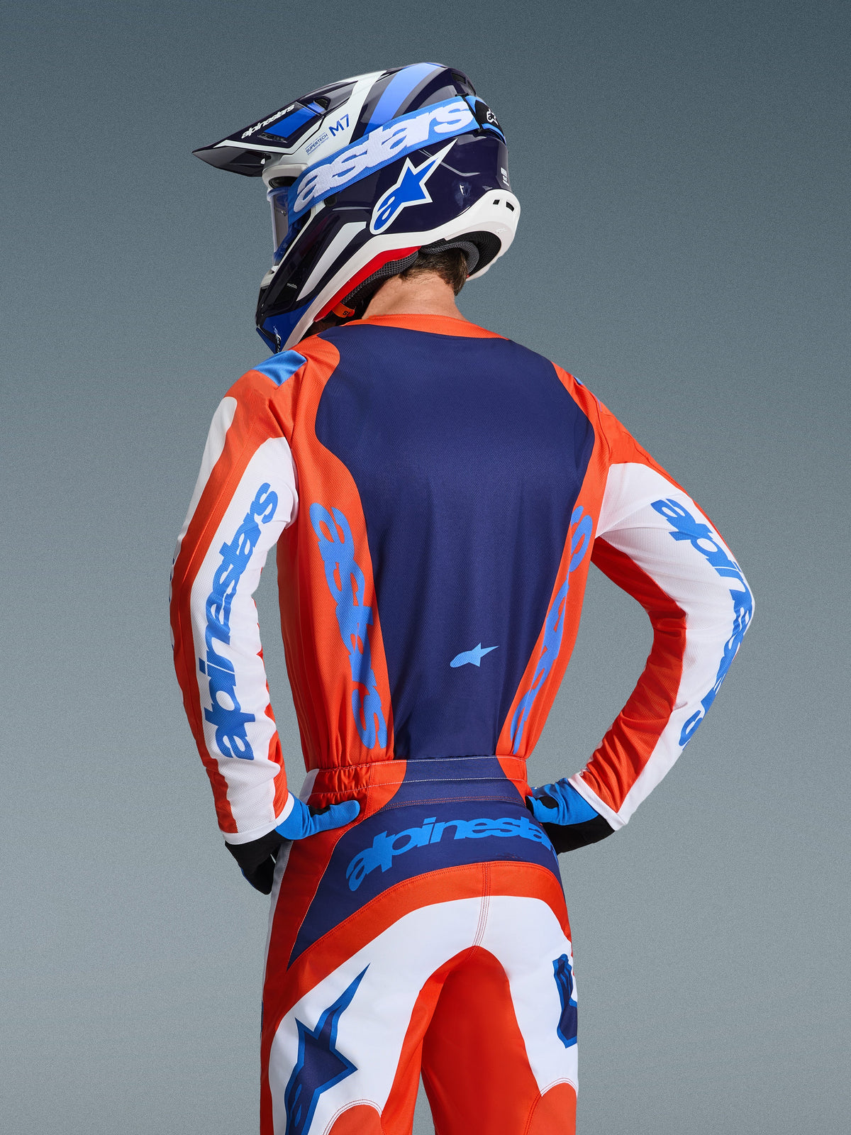 Alpinestars 2026 Fluid Grid Jersey, achteraanzicht van motorcrossrijder in oranje en UCLA Blue kledingset, donkerblauw rugpaneel, witte mouwaccenten met blauwe branding, bijpassende helm, hoogwaardige off-road kleding