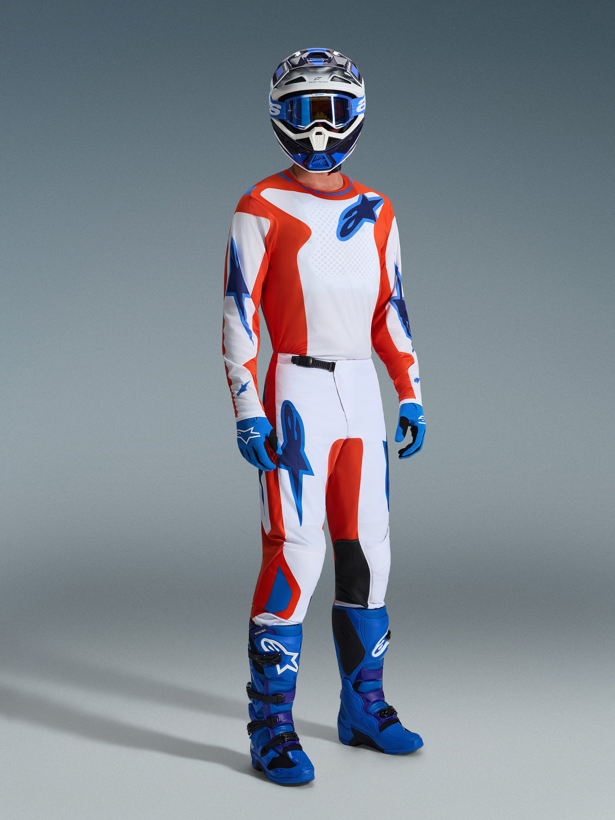 Motocrossrijder in de Alpinestars 2026 Fluid Grid Gear Set, witte jersey en broek met oranje en UCLA blauwe graphics, bijpassende blauwe helm en bril, blauwe handschoenen en felblauwe racelaarzen, gecoördineerde prestatie-outfit voor off-road rijden.
