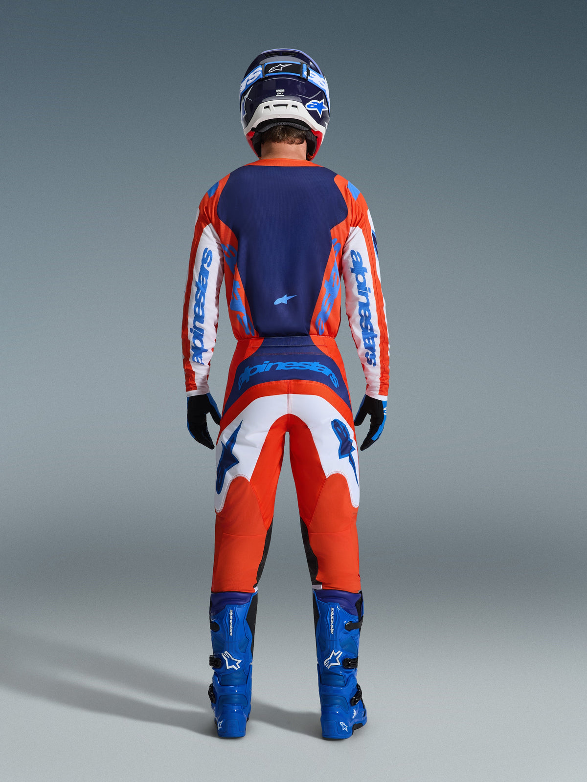 Alpinestars 2026 Fluid Grid Gear Set, motorcrosser van achteren gezien in een complete race-outfit in oranje en UCLA-blauw, jersey met lange mouwen met marineblauwe achterkant en witte mouwen, bijpassende oranje broek met blauwe graphics, blauwe laarzen en bijpassende helm, professionele off-road race-uitrusting.