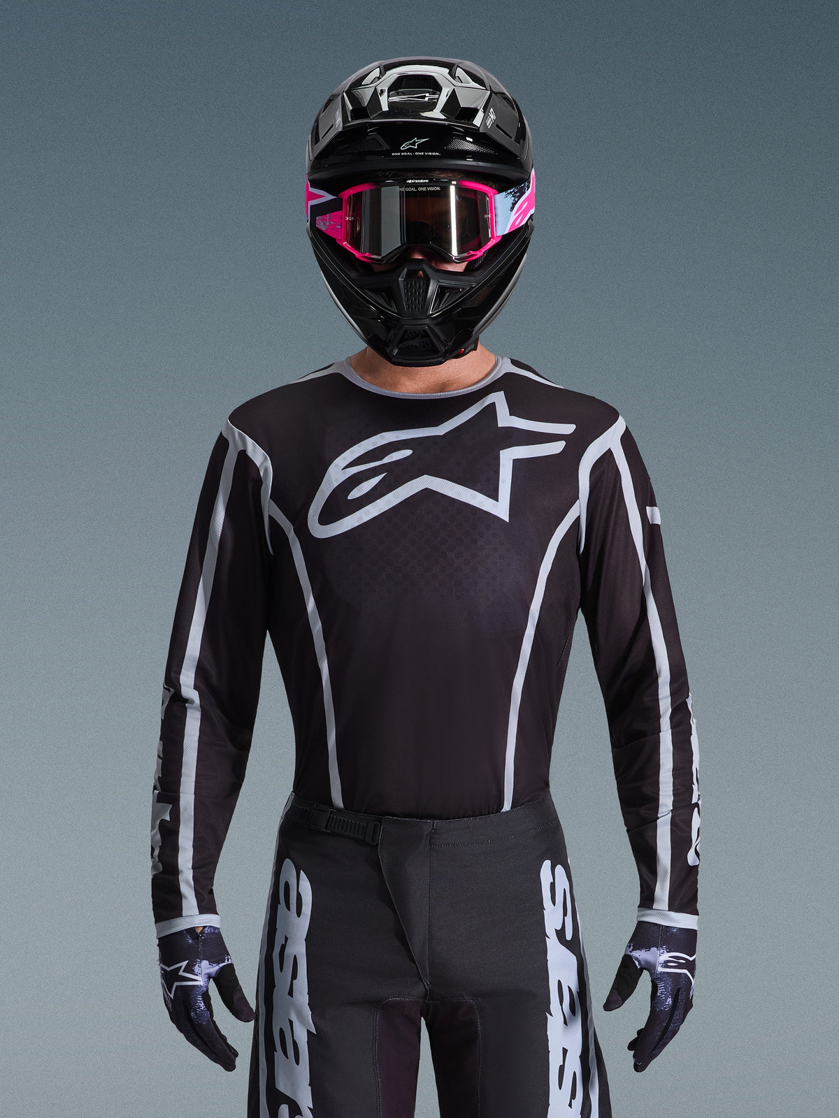 Alpinestars 2026 Fluid Apex kledingset, motorcross jersey en broek in zwart en wit, voorzien van een groot logo op de borst en geometrische lijnen, gedragen met een zwarte helm en roze bril, technische uitrusting voor off-road motorcross.