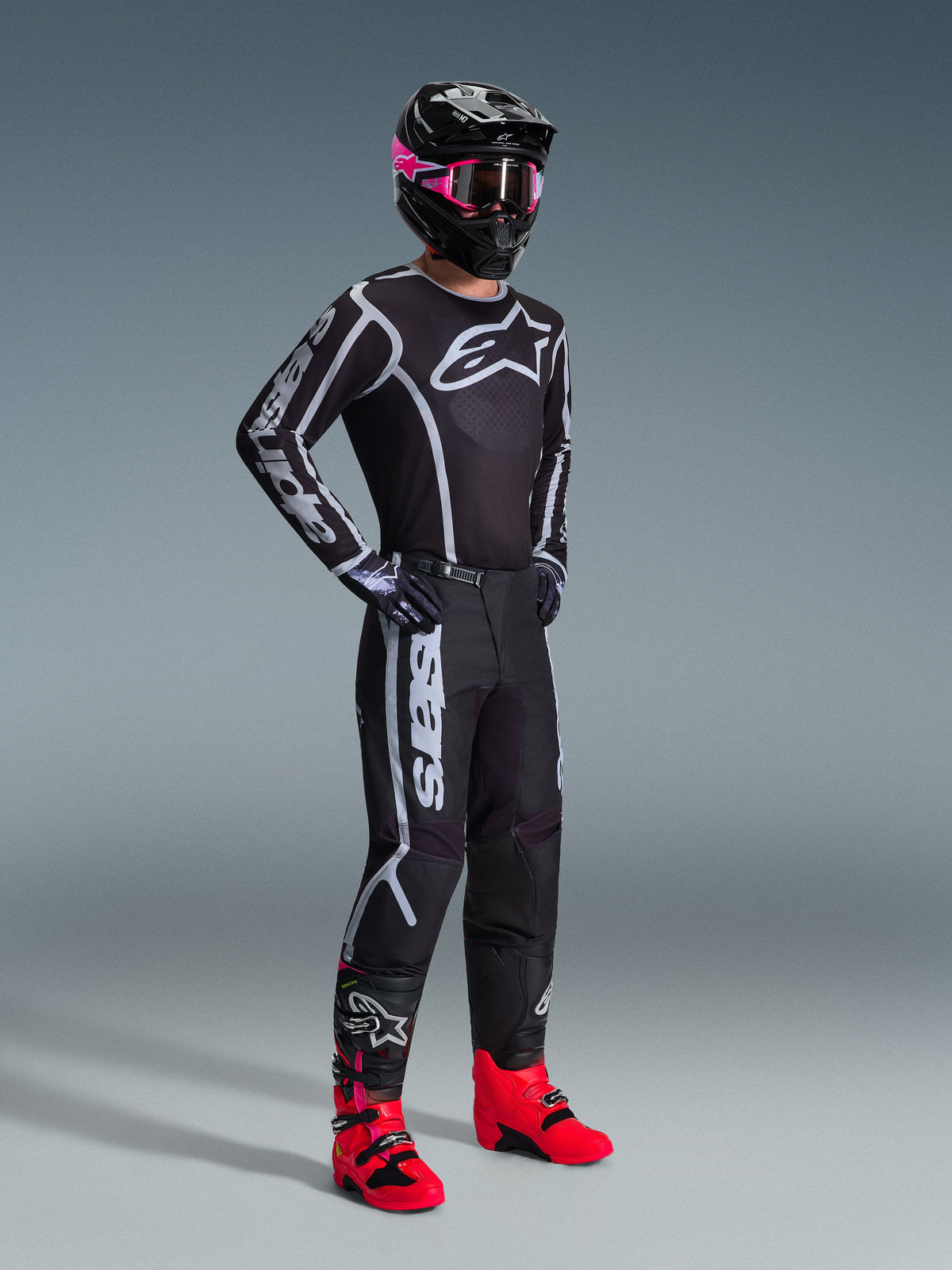 Alpinestars 2026 Fluid Apex uitrustingsset, motorcrosser in een zwarte jersey en broek met witte en grijze grafische accenten, zwarte helm met roze bril en felroze motorlaarzen, gecoördineerde professionele motorcross outfit