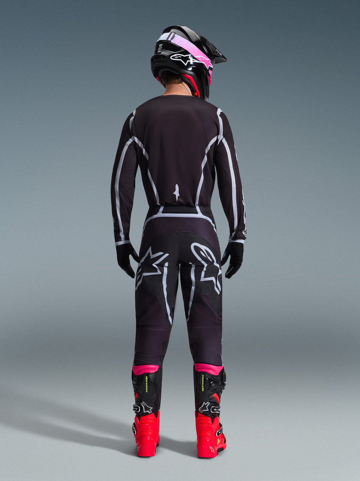 Motorcrosser van achteren gezien in de Alpinestars 2026 Fluid Apex Gear Set, zwarte jersey en broek met grijze geometrische lijnen en sterlogo's, zwarte helm met roze accenten, felrode en zwarte laarzen, gecoördineerde motorcross race-uitrusting
