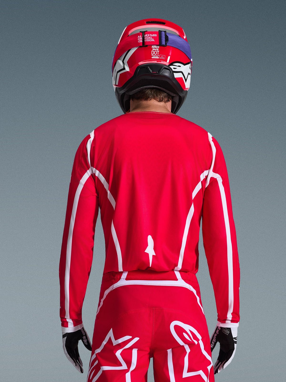 Alpinestars 2026 Fluid Apex Jersey, motorcross shirt, rood en wit, achteraanzicht van een motorcrosser met geometrische witte patronen, bijpassende motorcrossbroek en rode helm, complete race-uitrusting voor off-road prestaties