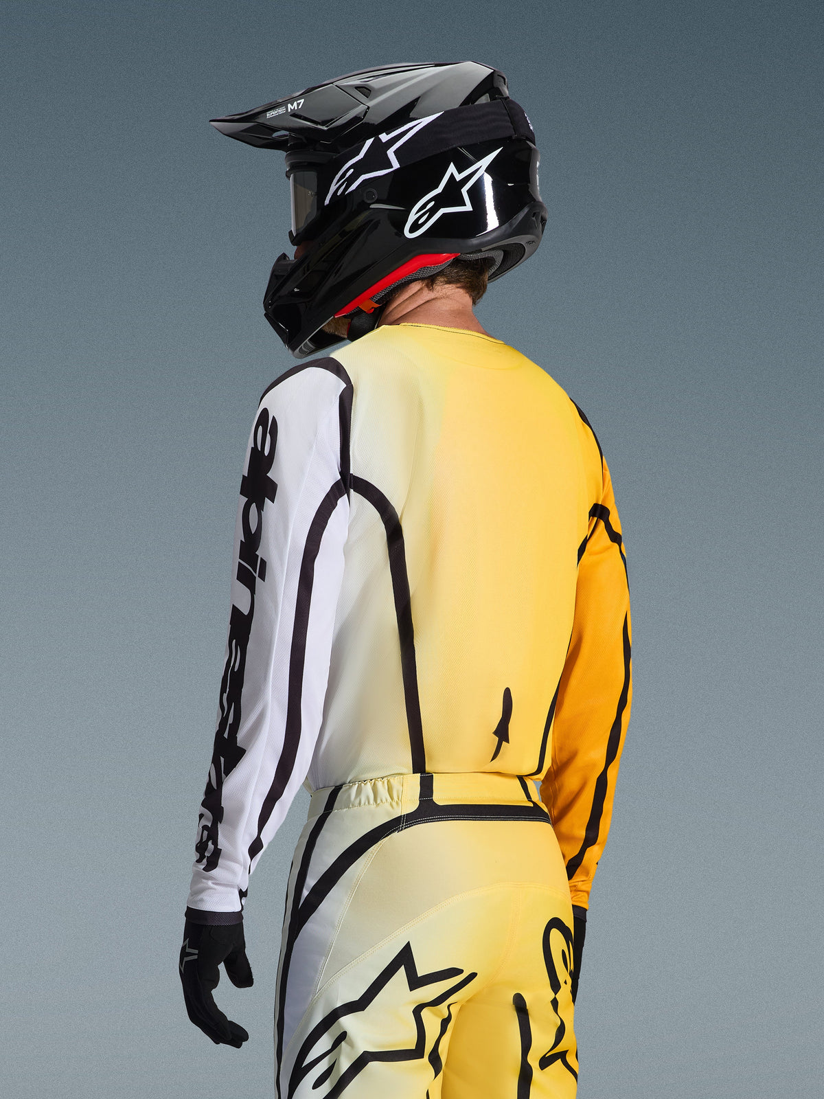 Driekwart achteraanzicht van een motorcrosser die de 2026 Fluid Apex Jersey in Yellow Black draagt, met een kleurverloop van geel naar wit, opvallende zwarte Alpinestars-branding op de mouw, gedragen met een bijpassende broek en een zwarte motorcrosshelm.