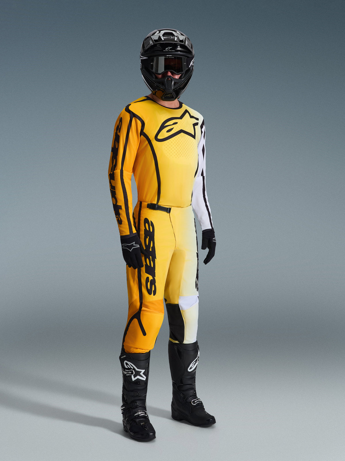 Motocrossrijder in de Alpinestars 2026 Fluid Apex Jersey in geel en zwart, met een kleurverloop van geel naar wit en een groot zwart logo op de borst, gecombineerd met bijpassende broek, zwarte helm en zwarte laarzen, complete race-uitrusting voor off-road motorcross.